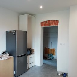 Wojtek Turowski - Wnętrze kuchni w trakcie wykańczania, widoczna lodówka, zabudowa meblowa i przejście z łukiem wykończonym czerwoną cegłą do innego pomieszczenia, na podłodze szare płytki.