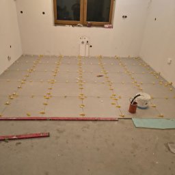 Wojtek Turowski - Układanie szarych płytek podłogowych w jasnym pomieszczeniu. Widoczne poziomice, krzyżyki dystansowe i wiadro z narzędziami. Okno w tle.