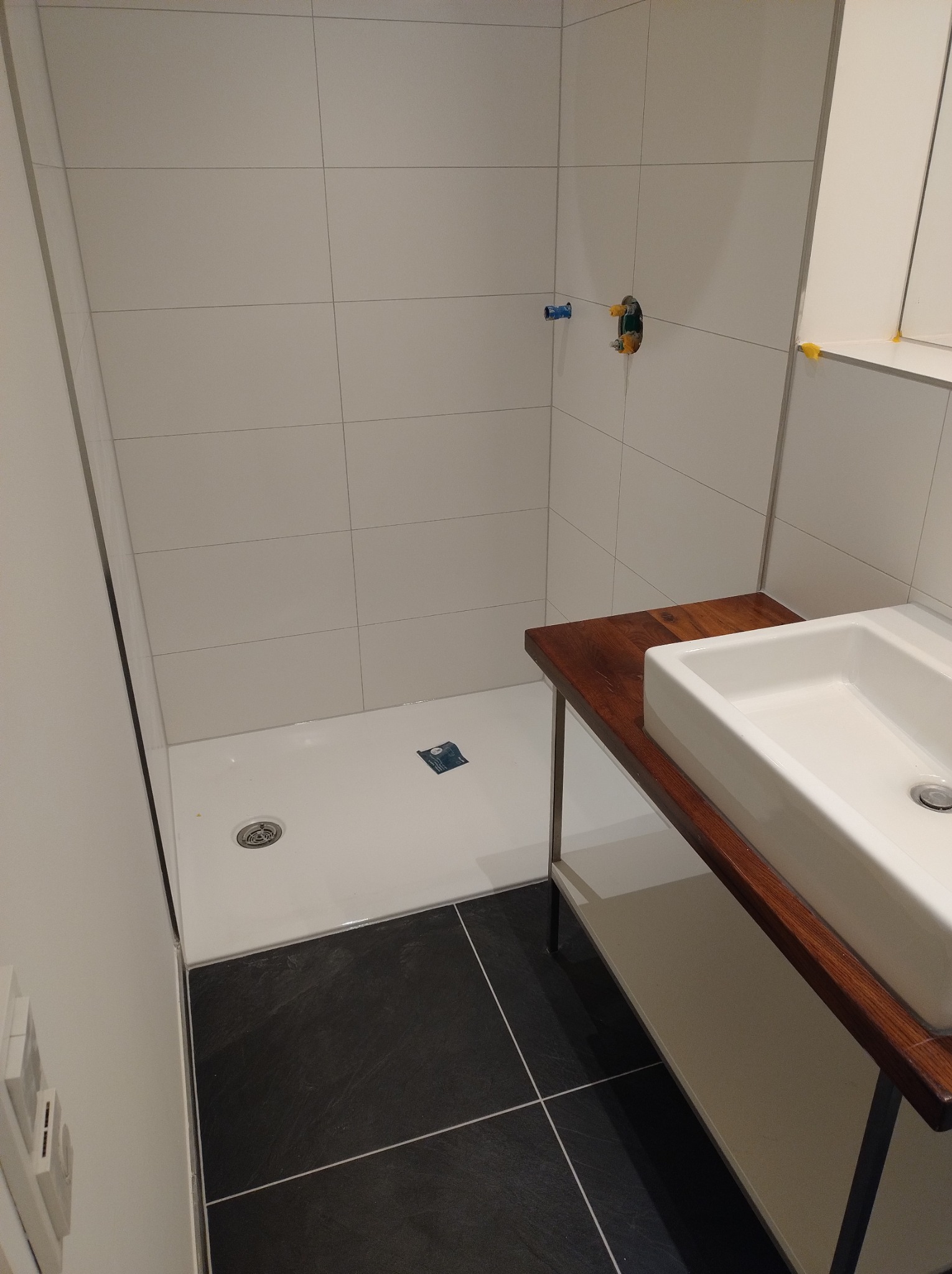 Nowoczesna łazienka z białymi płytkami, czarną podłogą i drewnianym blatem pod umywalką, widoczne niepodłączone zawory prysznicowe.