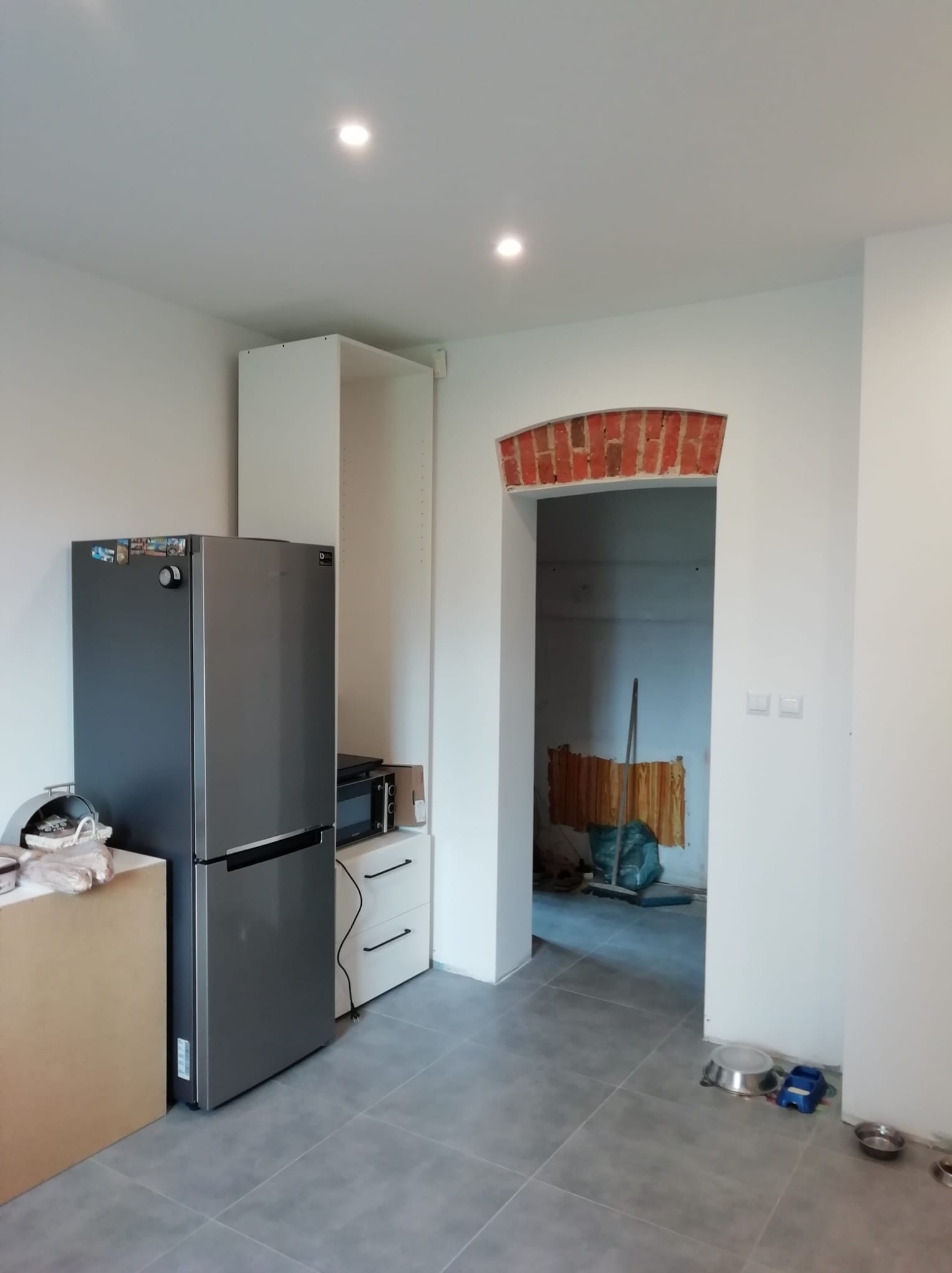 Wnętrze kuchni w trakcie wykańczania, widoczna lodówka, zabudowa meblowa i przejście z łukiem wykończonym czerwoną cegłą do innego pomieszczenia, na podłodze szare płytki.