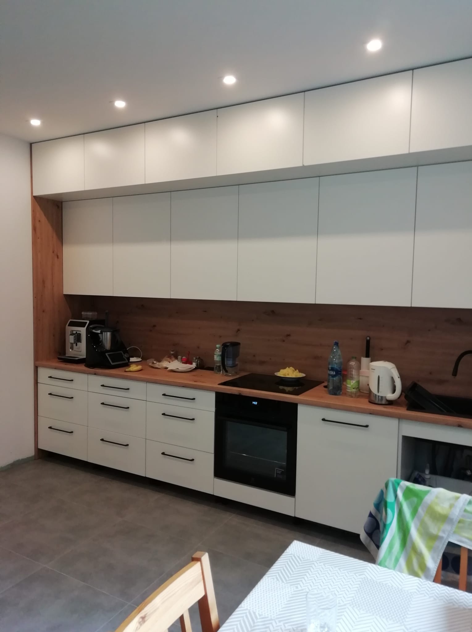 Nowoczesna kuchnia z białymi szafkami, drewnianym panelem, czarnymi uchwytami i sprzętem AGD na drewnianym blacie.