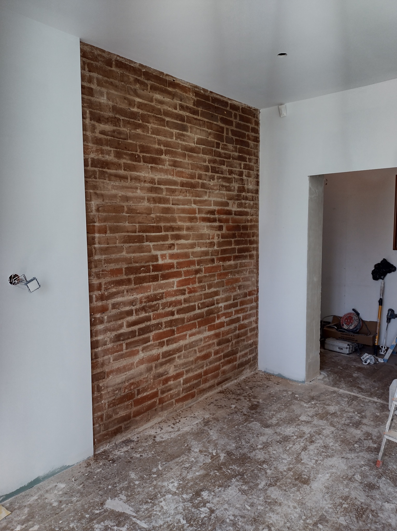 Fragment ściany z cegły klinkierowej w trakcie prac wykończeniowych, widoczne ślady zaprawy na podłodze i białe ściany, brak listew przypodłogowych, przygotowane gniazdko elektryczne.