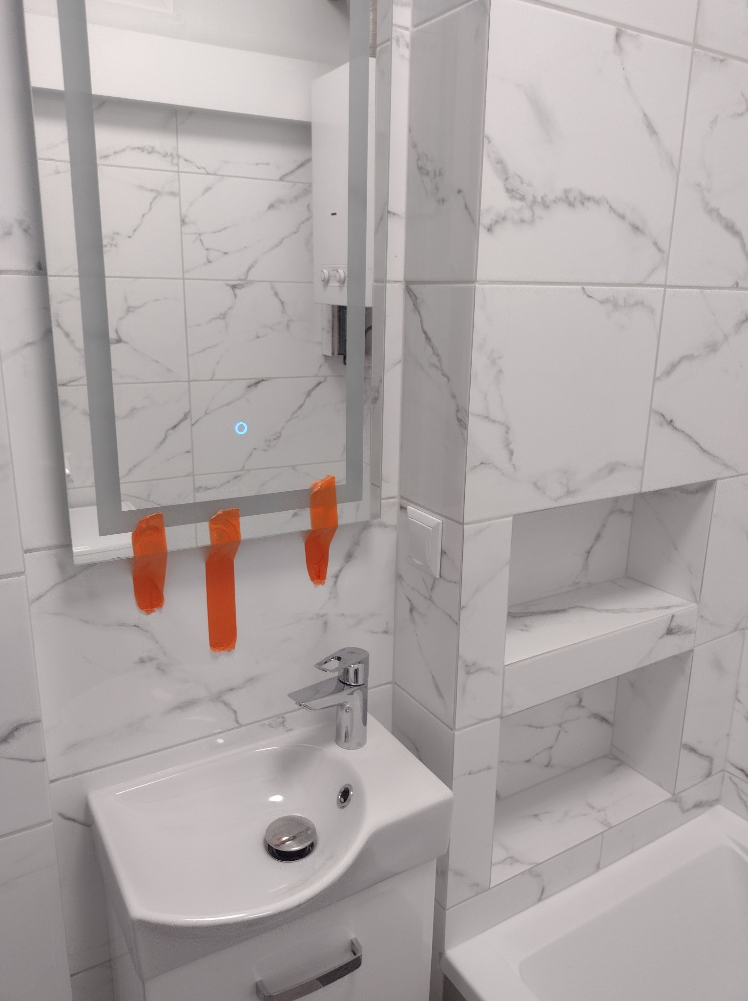 Nowoczesna łazienka z marmurowymi płytkami, umywalką z szafką, podświetlanym lustrem zabezpieczonym pomarańczową taśmą i wnękami w ścianie.