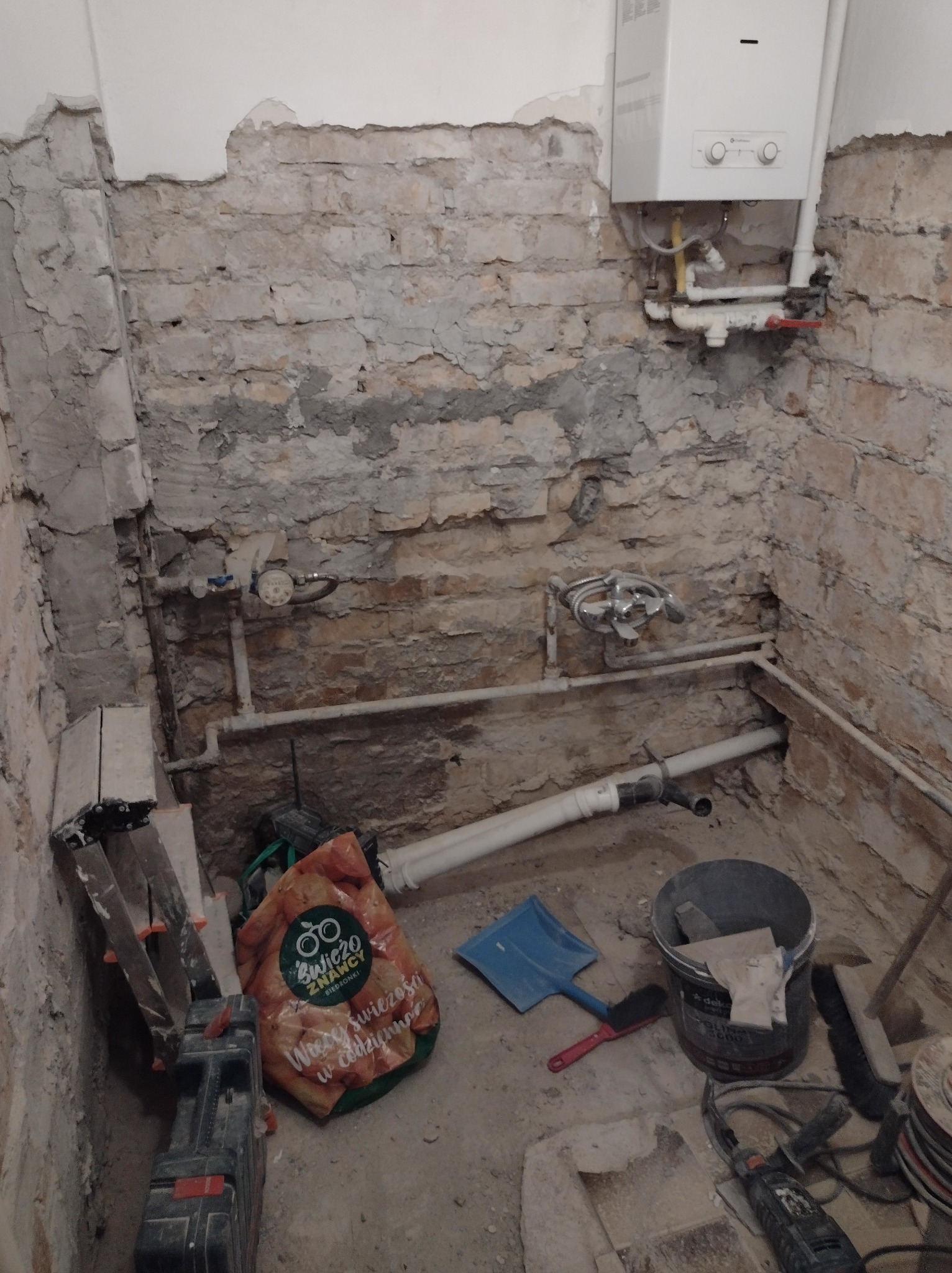 Łazienka w trakcie demontażu płytek, odsłonięta cegła, widoczne instalacje wodne i piec gazowy, narzędzia i wiadro na podłodze.