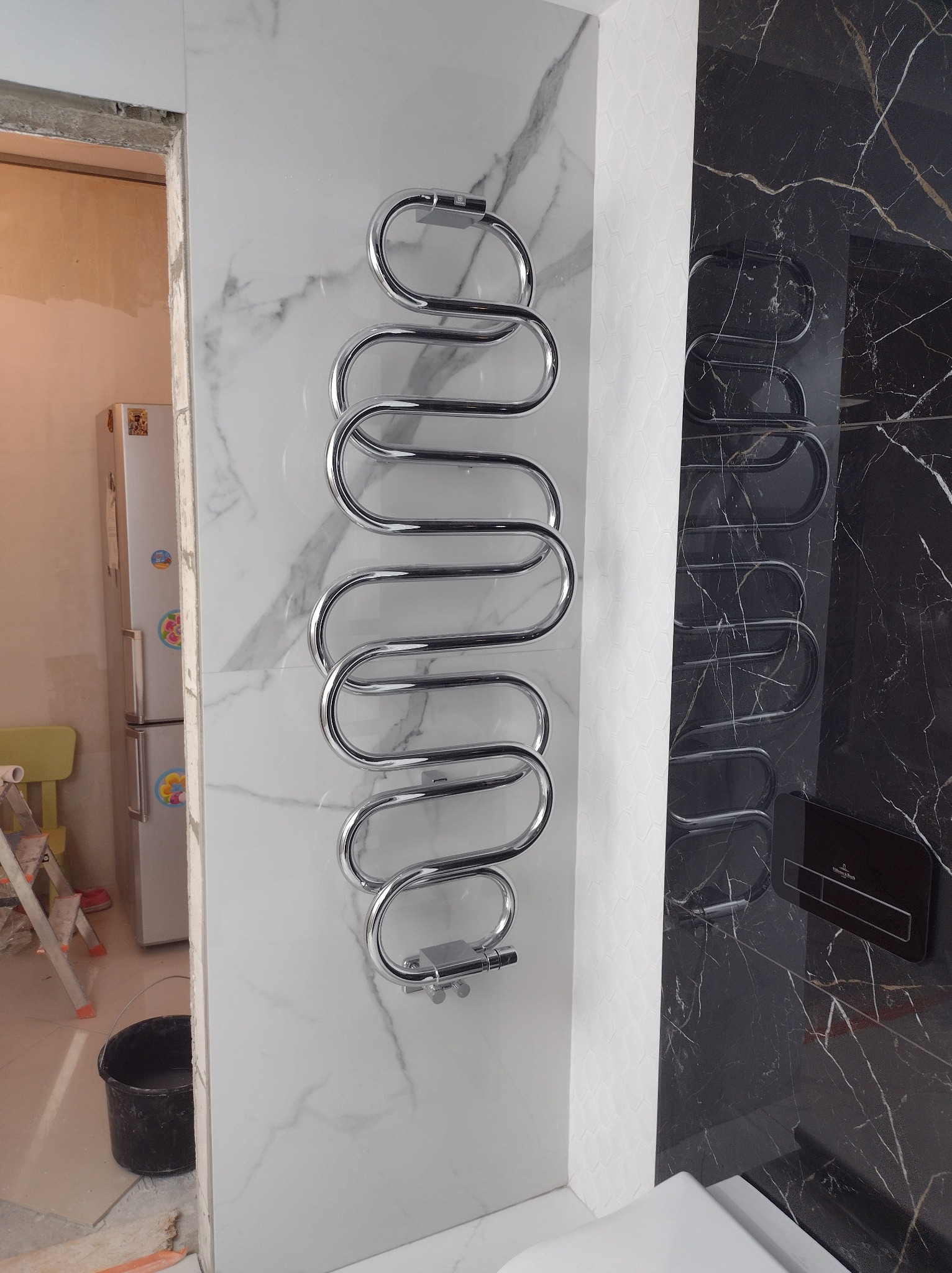 Chromowany grzejnik łazienkowy w kształcie węża na białej marmurowej ścianie, obok czarnej marmurowej ściany z widocznym przyciskiem spłuczki Villeroy & Boch, w tle fragment łazienki w trakcie prac...