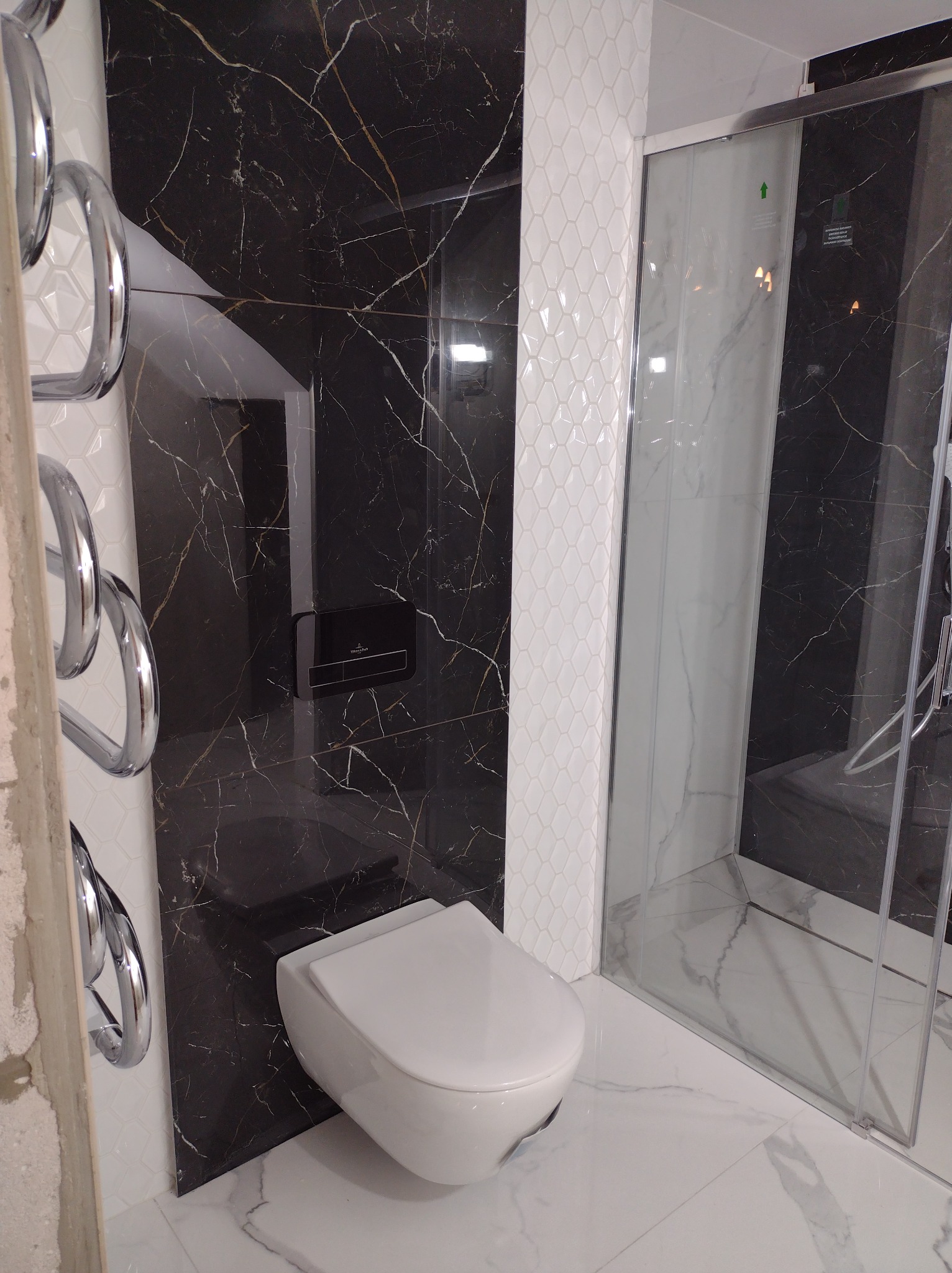 Nowoczesna łazienka z czarnymi płytkami imitującymi marmur, białą mozaiką na ścianie, podwieszaną toaletą i szklaną kabiną prysznicową.