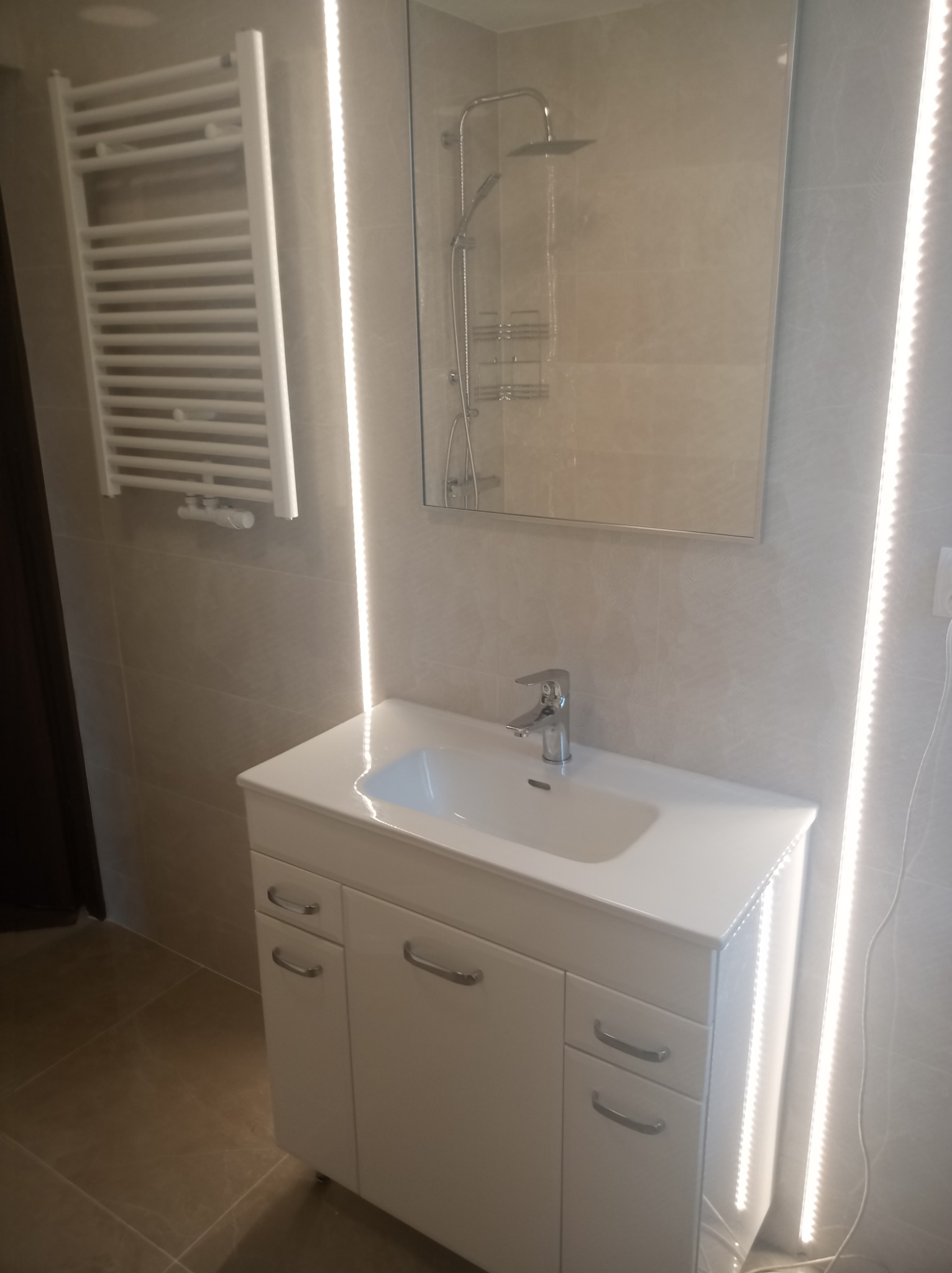 Nowoczesna łazienka z białymi meblami łazienkowymi, umywalką, grzejnikiem drabinkowym i podświetlanym lustrem z kabiną prysznicową.