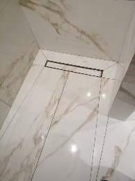 Białe, marmurkowe płytki podłogowe w łazience z czarnym odpływem liniowym, widoczne delikatne spoiny.