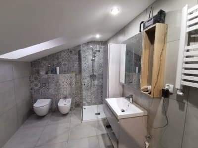 Nowoczesna łazienka na poddaszu ze skośnym sufitem, szara mozaika na ścianie, kabina prysznicowa typu walk-in, umywalka z szafką w kolorze taupe i grzejnik drabinkowy.