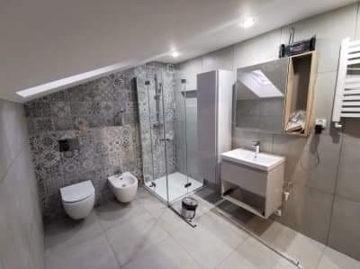 Nowoczesna łazienka na poddaszu z kabiną prysznicową ze szklanymi drzwiami, umywalką z szafką, toaletą i bidetem, wykończona szarymi płytkami z dekoracyjnym wzorem na ścianie za toaletą i bidetem.