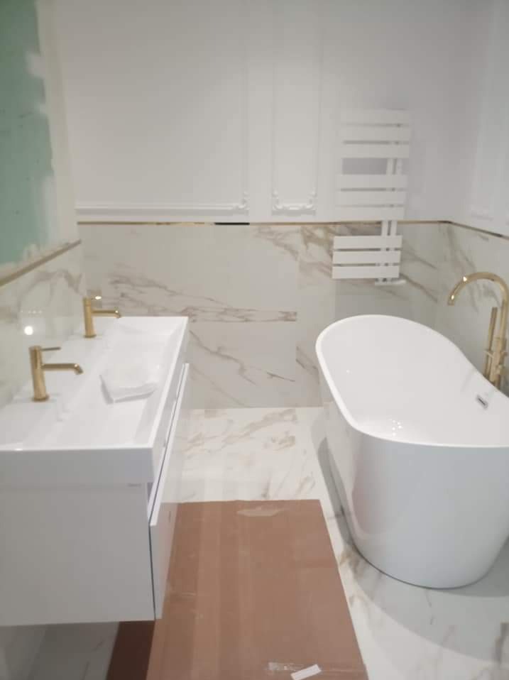 Nowoczesna łazienka w trakcie remontu: biała wanna wolnostojąca, umywalka z szafką i złotą armaturą na tle marmurowych płytek z delikatnym złotym akcentem, widoczny grzejnik drabinkowy i fragment...