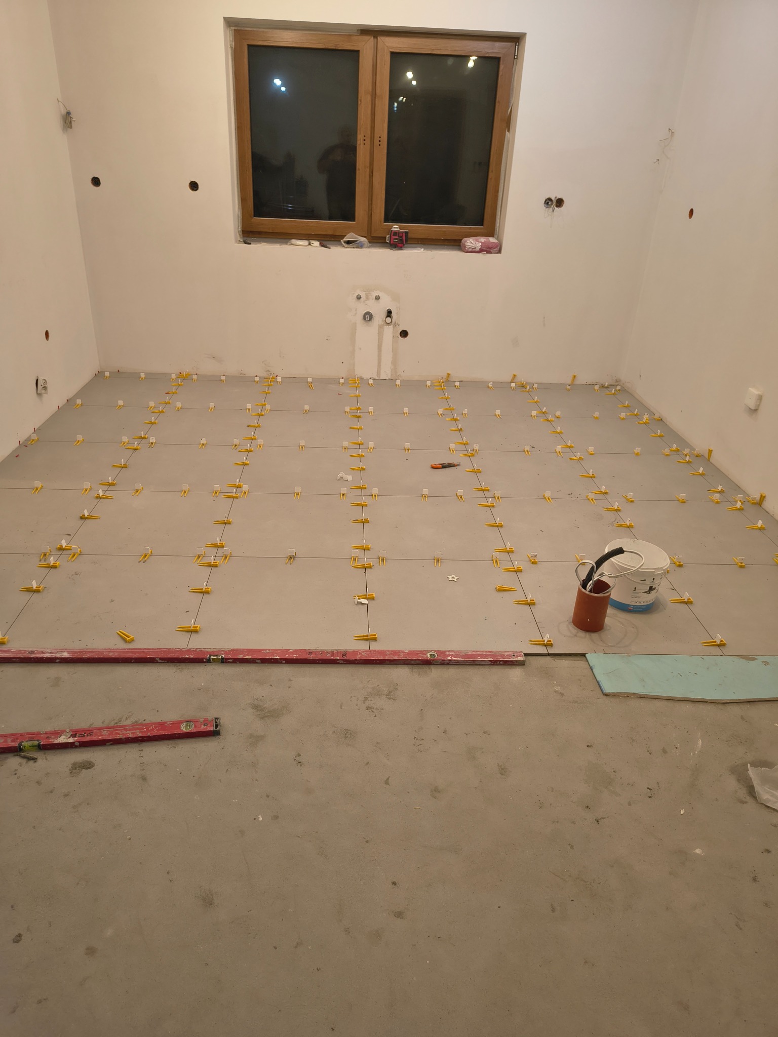 Układanie szarych płytek podłogowych w jasnym pomieszczeniu. Widoczne poziomice, krzyżyki dystansowe i wiadro z narzędziami. Okno w tle.