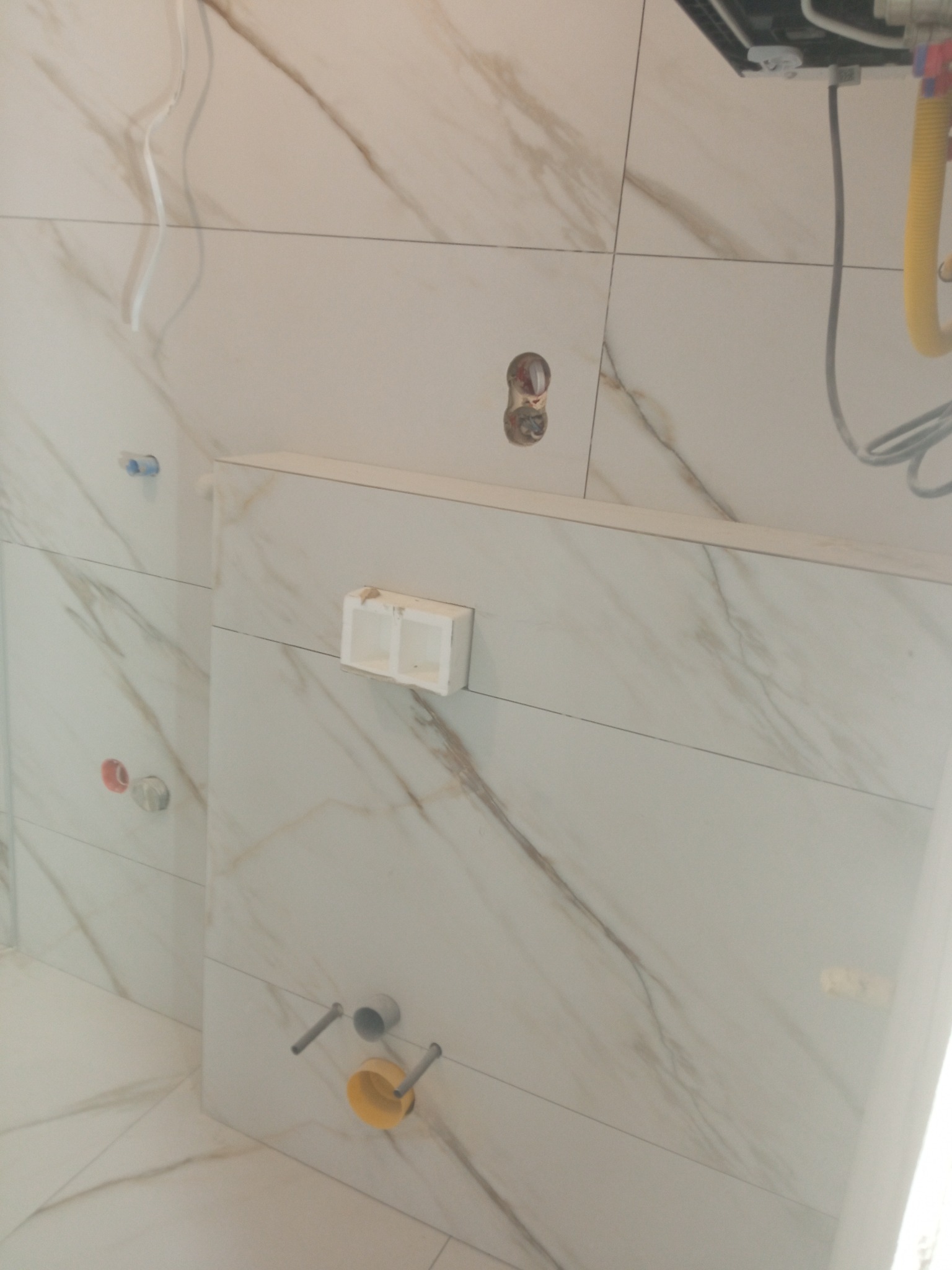 Białe płytki imitujące marmur z widocznymi instalacjami hydraulicznymi i elektrycznymi w trakcie wykańczania łazienki. Widoczne otwory i puszki instalacyjne.