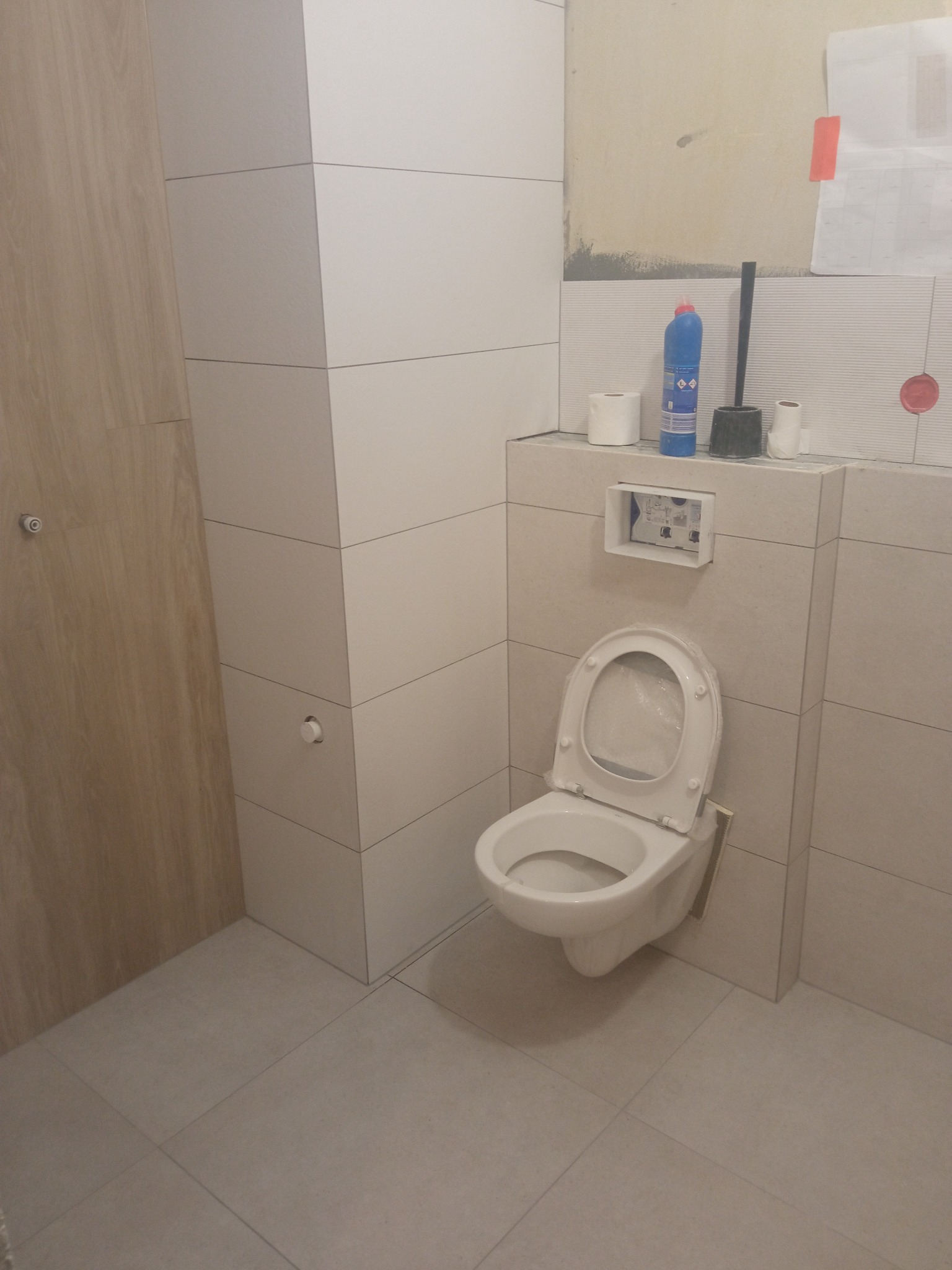 Nowoczesna toaleta podwieszana w trakcie montażu, obudowana płytkami w odcieniach bieli i beżu. Widoczne akcesoria instalacyjne i fragment ściany w trakcie wykończenia.