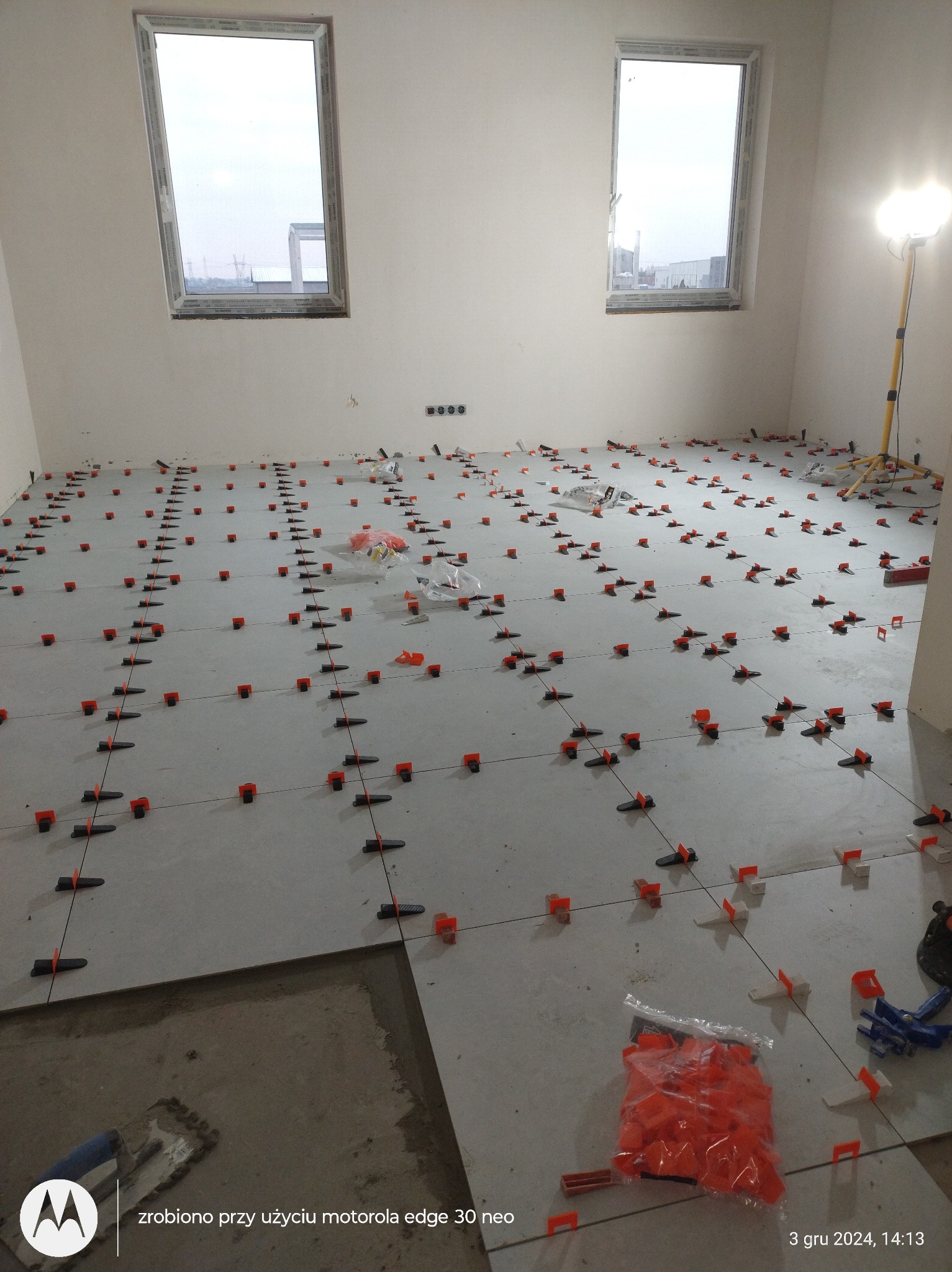 Układanie jasnych płytek podłogowych z użyciem systemu poziomowania. Widoczne krzyżyki dystansowe i kliny. W tle dwa okna i lampa budowlana. Etap prac wykończeniowych.