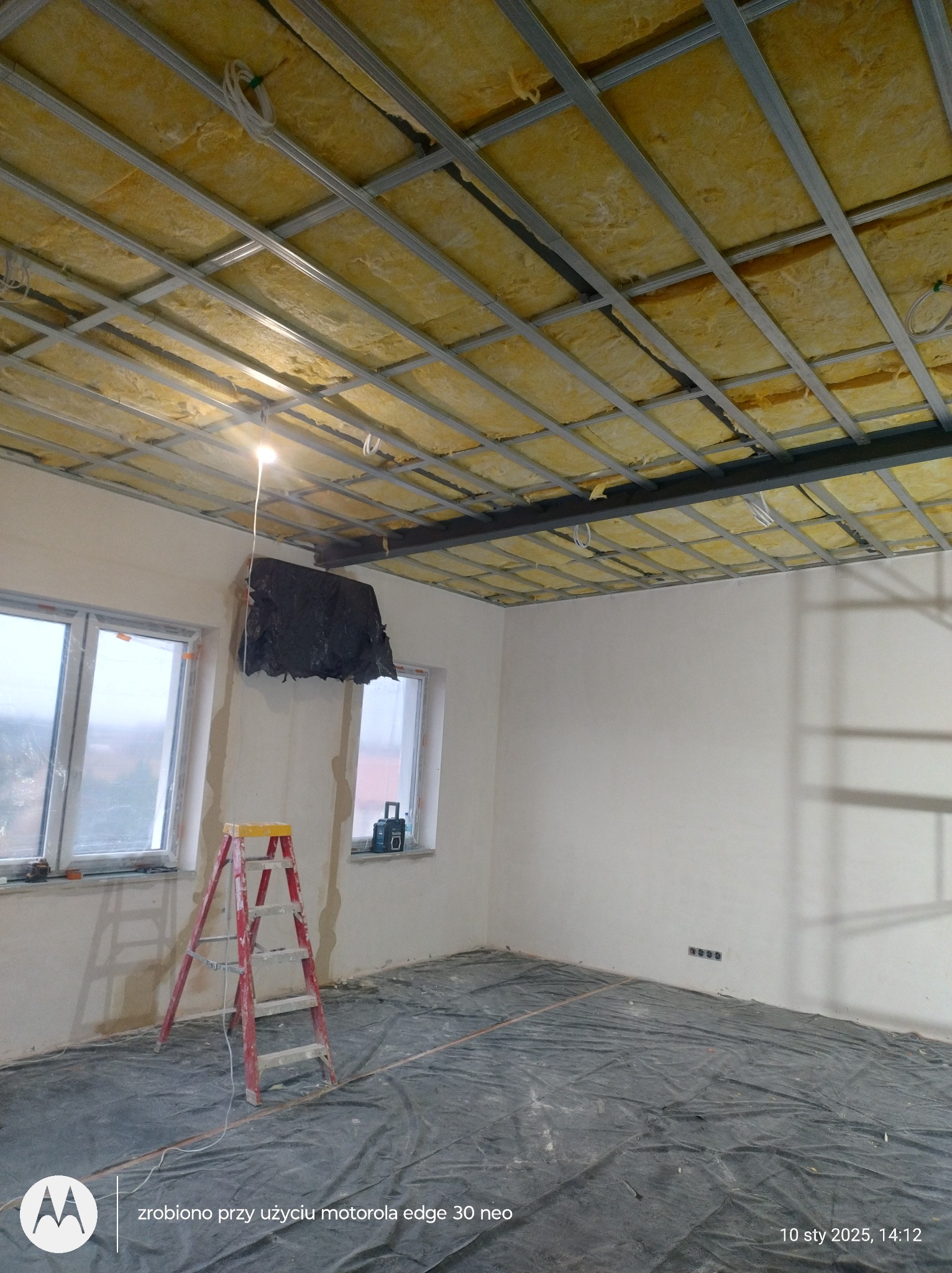 Montaż sufitu podwieszanego z izolacją z wełny mineralnej. Widoczne profile, izolacja, drabina i zabezpieczona folią podłoga. Prace wykończeniowe w pomieszczeniu.