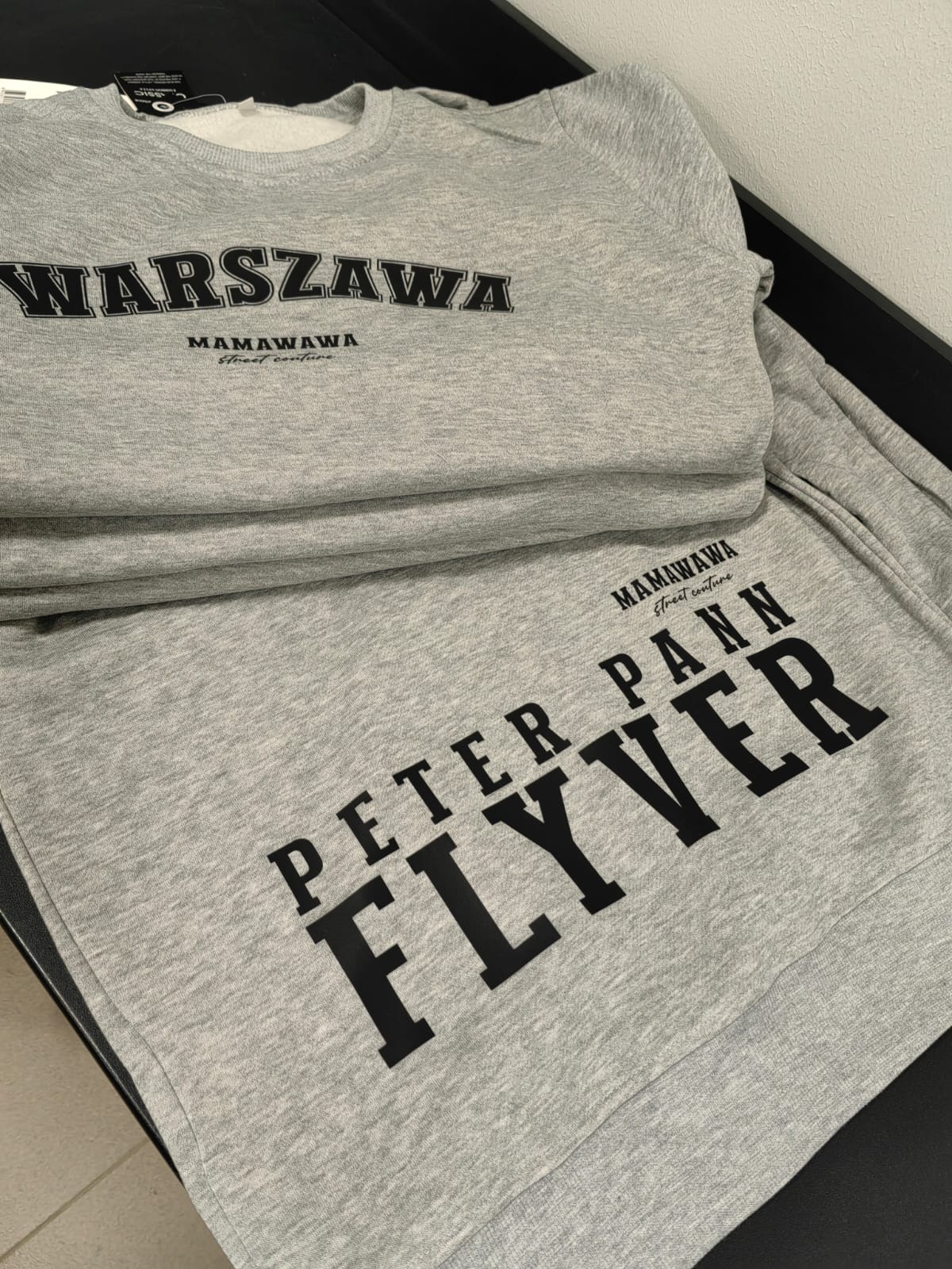 Stos szarych bluz z czarnymi nadrukami 'WARSZAWA' i 'PETER PANN FLYVER' oraz logotypem 'MAMAWAVA Street couture'.
