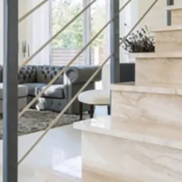 Jasne marmurowe schody z metalową balustradą w nowoczesnym wnętrzu z szarym pikowanym narożnikiem i białym krzesłem w tle.