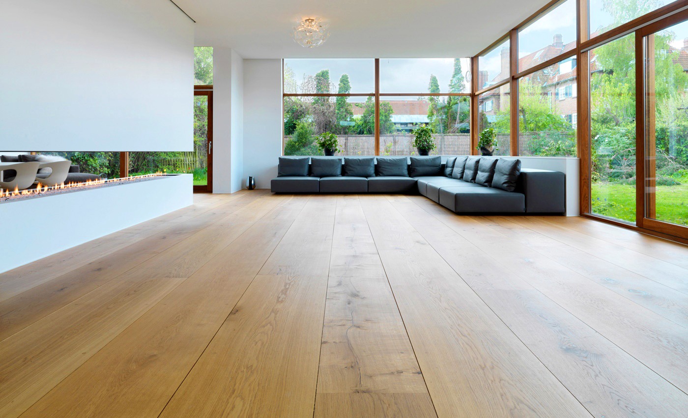Jasna, drewniana podłoga w przestronnym salonie z dużymi oknami, minimalistycznym kominkiem i szarą sofą modułową.