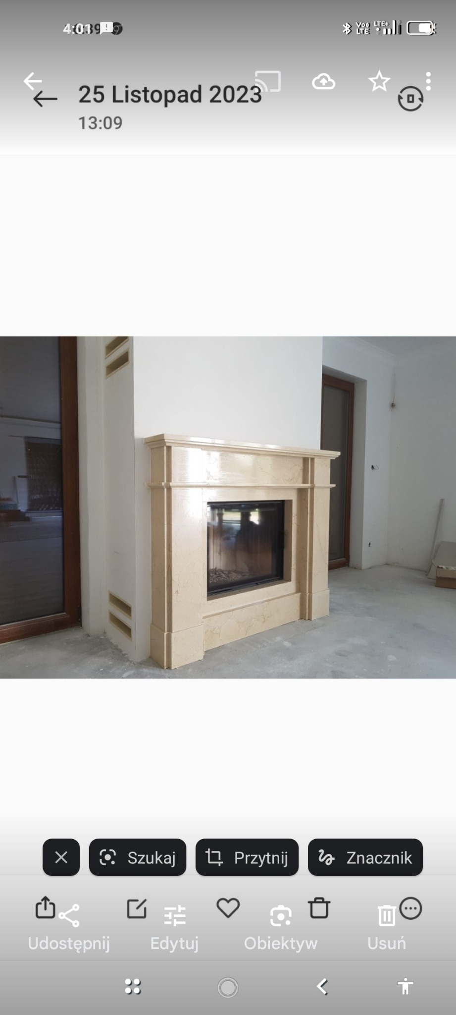 Marmurowy kominek z czarnym wkładem w surowym wnętrzu, widoczne niedokończone wykończenia ścian i podłogi, obok drzwi balkonowe.