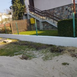 AXVI Usługi Ogólnoremontowe Julia Tubiak - Świeżo pomalowany na biało murek oporowy z widocznymi śladami ziemi, ogrodzenie z siatki i drewnianych sztachet w tle, trawnik i częściowo odsłonięta ziemia w pierwszym planie.