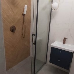 AXVI Usługi Ogólnoremontowe Julia Tubiak - Nowoczesna łazienka z kabiną prysznicową z czarnymi ramami, drewnopodobnymi płytkami na ścianie prysznica, armaturą w kolorze miedzi, wiszącą szafką z umywalką i designerską lampą sufitową.