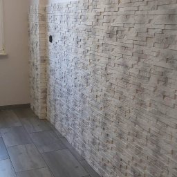 AXVI Usługi Ogólnoremontowe Julia Tubiak - Ściana wyłożona dekoracyjną, jasną imitacją kamienia, z widocznym otworem na gniazdko elektryczne, połączona z szarą podłogą z płytek imitujących drewno.