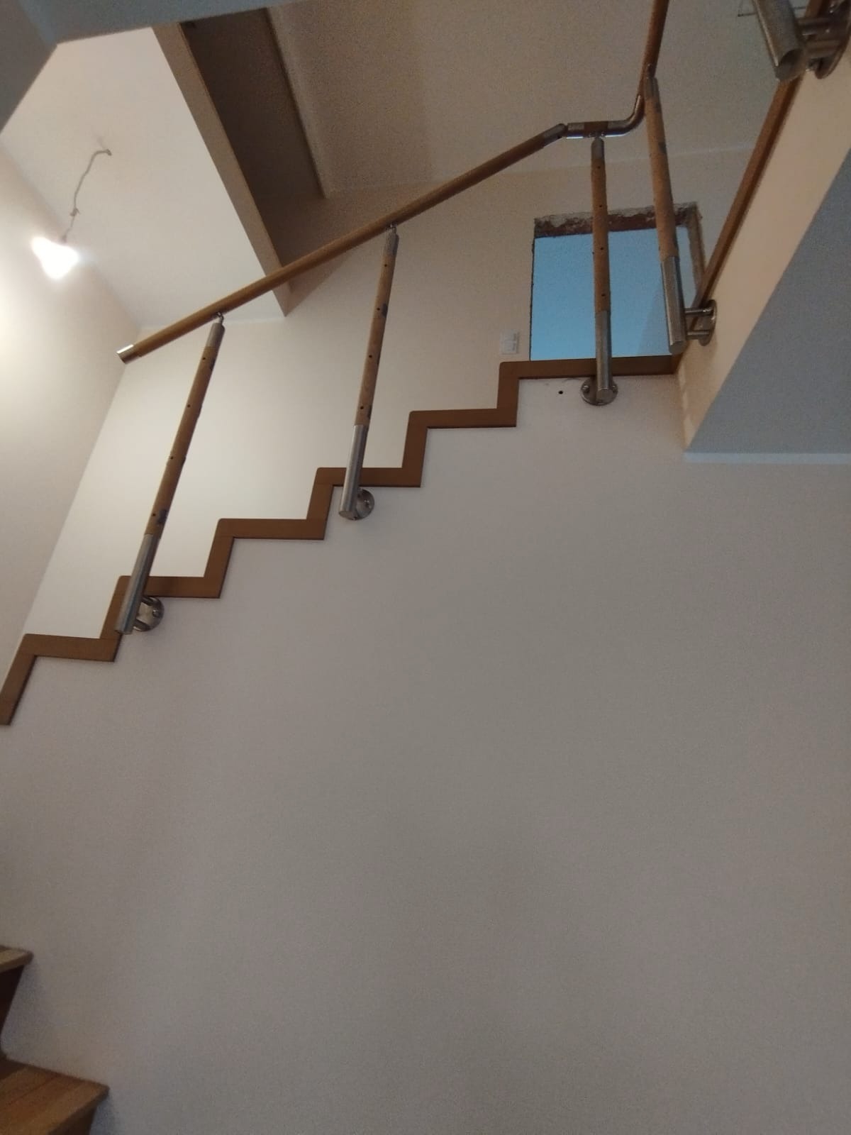 Schody z drewnianą poręczą i metalowymi wspornikami przymocowanymi do ściany w kształcie zygzaka, widoczne oświetlenie sufitowe.