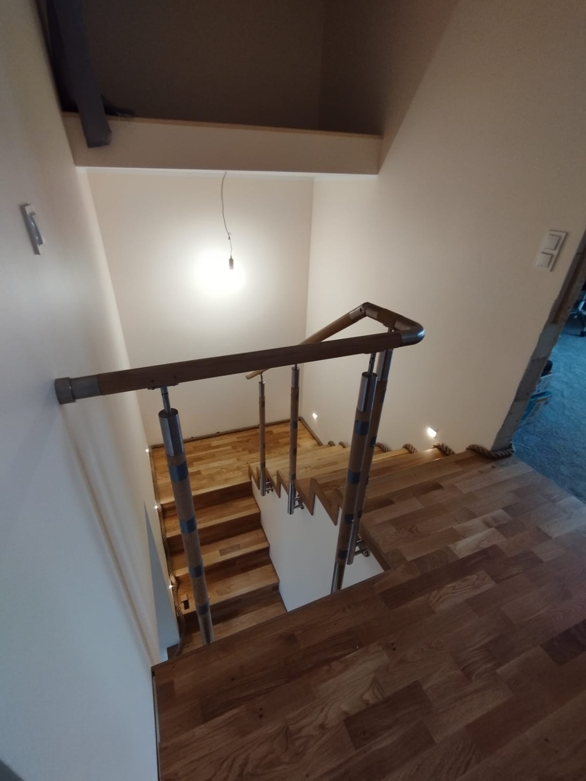 Drewniane schody z metalowymi balustradami, oświetlone małymi punktowymi lampkami w ścianie, widok z góry.