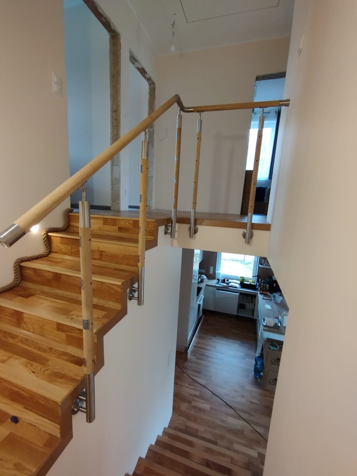 Drewniane schody z balustradą z elementami ze stali nierdzewnej i ozdobnym sznurem. W tle widoczne drzwi w stanie surowym i wnętrze kuchni.