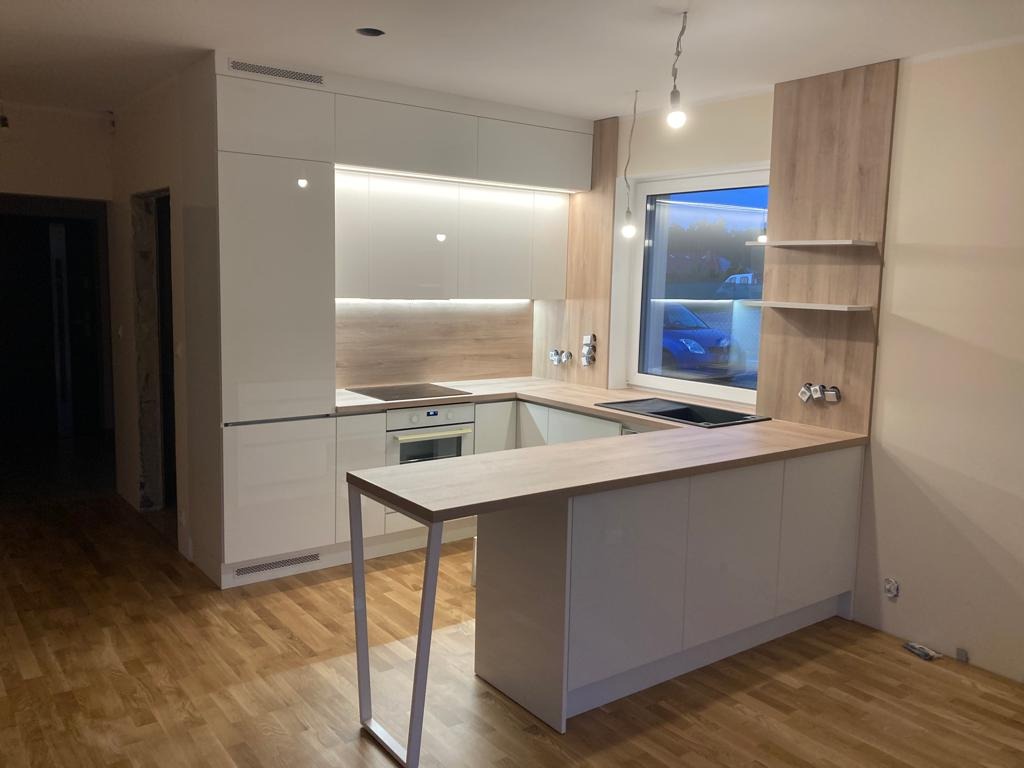 Nowoczesna kuchnia z białymi szafkami i drewnianym blatem, oświetlona wiszącymi lampami, widok na okno z niebieskim samochodem na zewnątrz, drewniana podłoga.