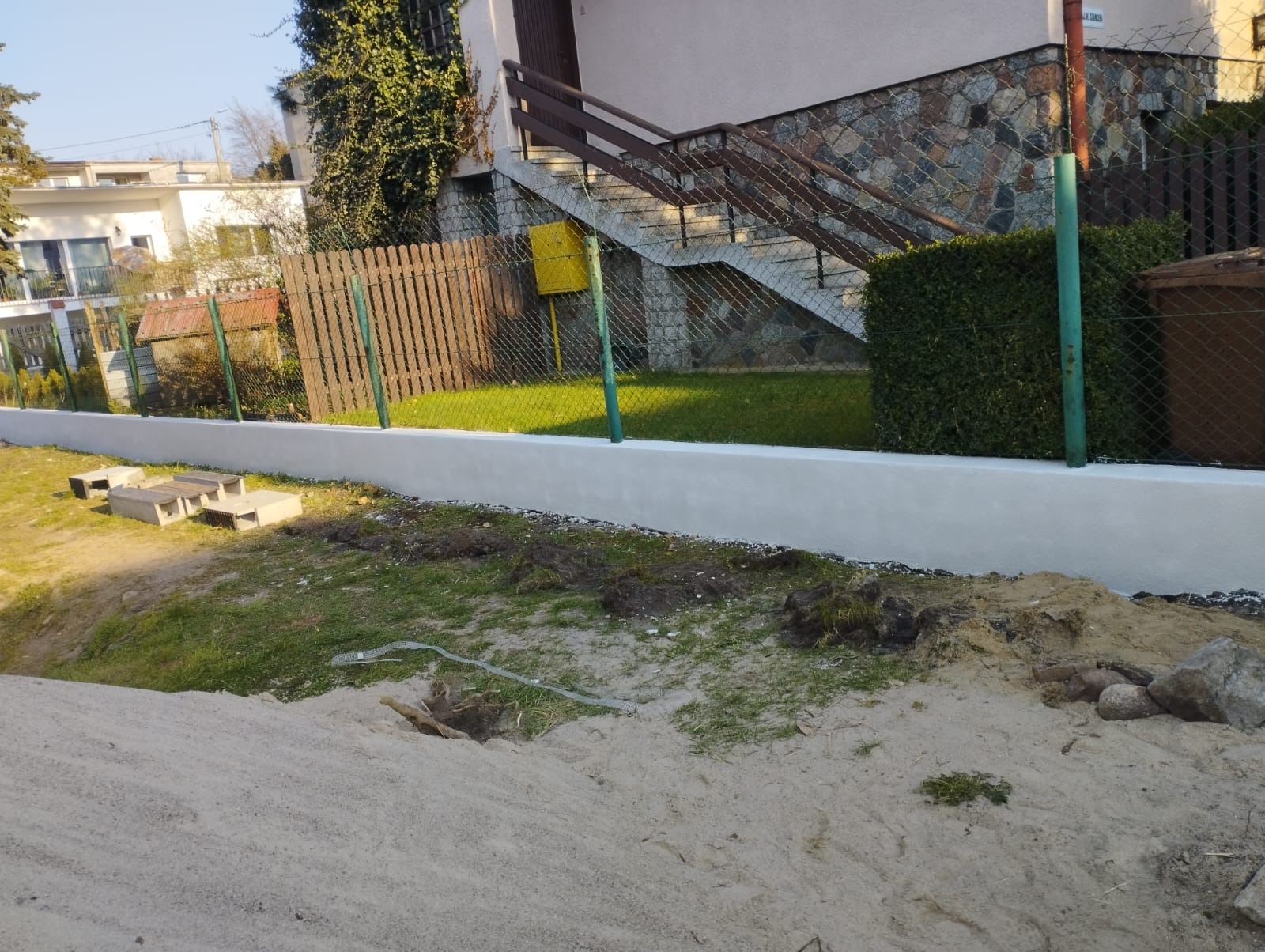 Świeżo pomalowany na biało murek oporowy z widocznymi śladami ziemi, ogrodzenie z siatki i drewnianych sztachet w tle, trawnik i częściowo odsłonięta ziemia w pierwszym planie.