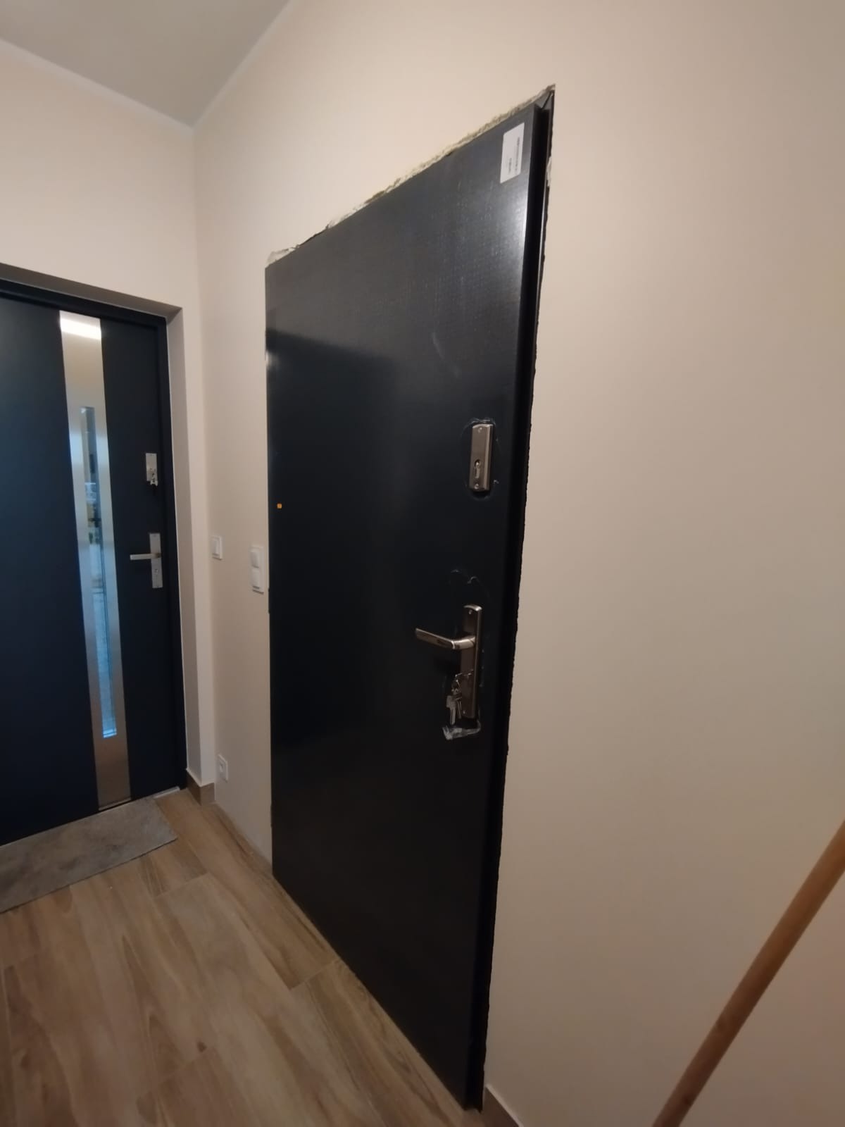 Ciemne drzwi wejściowe z srebrną klamką i kluczami, widoczne w korytarzu z drewnopodobną podłogą i drugimi drzwiami w tle, przed wykończeniem ościeżnicy.