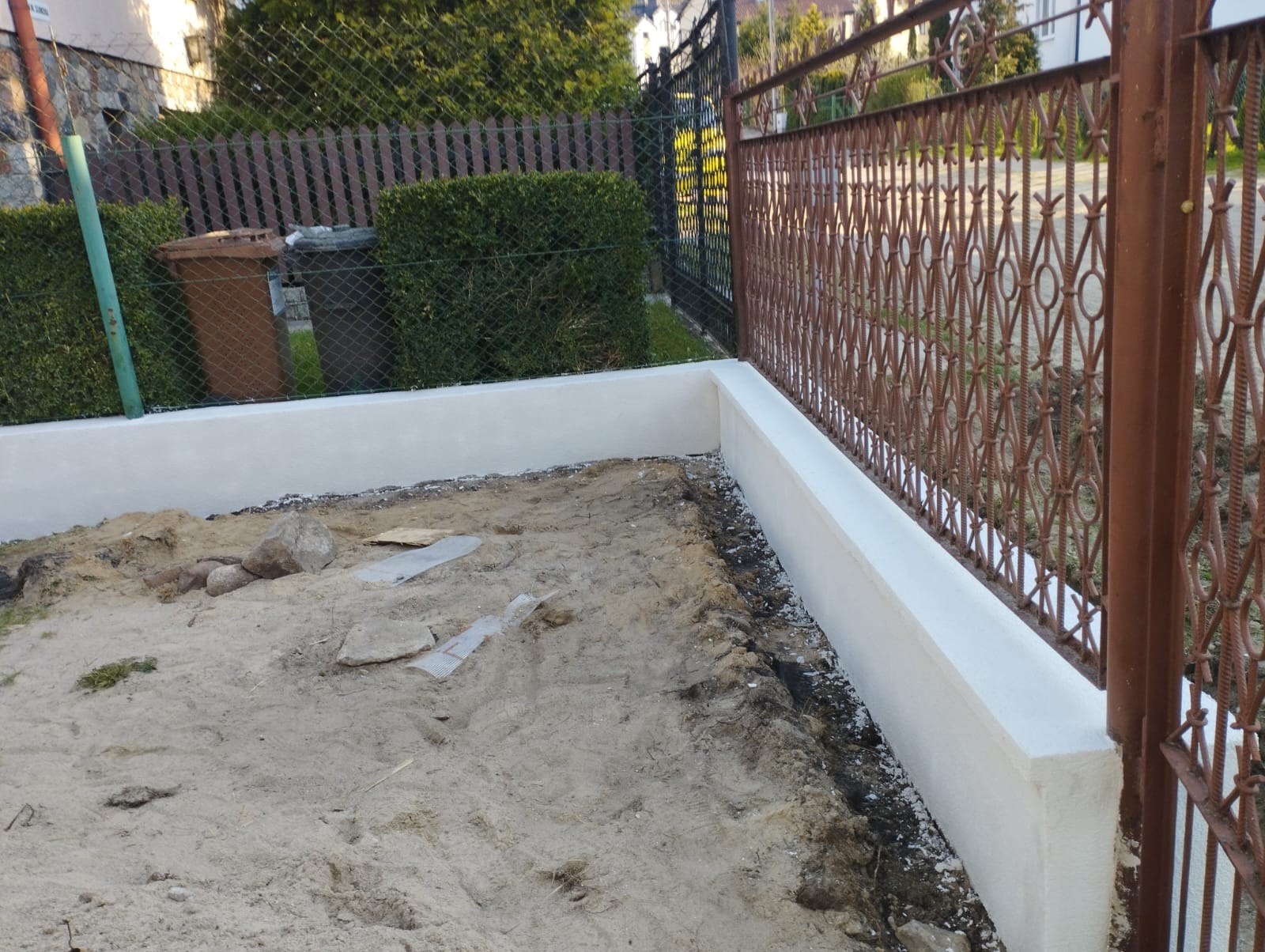 Świeżo otynkowana podmurówka ogrodzenia z metalową, ozdobną kratą, widoczne ślady prac ziemnych i piasek.