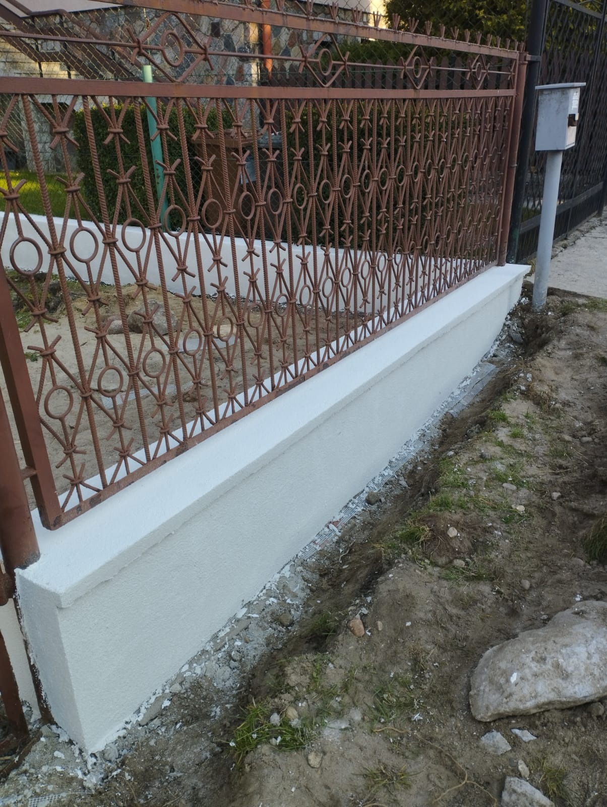 Świeżo otynkowany cokół ogrodzenia z metalową, ozdobną kratą, widoczne ślady prac wykończeniowych na gruncie.