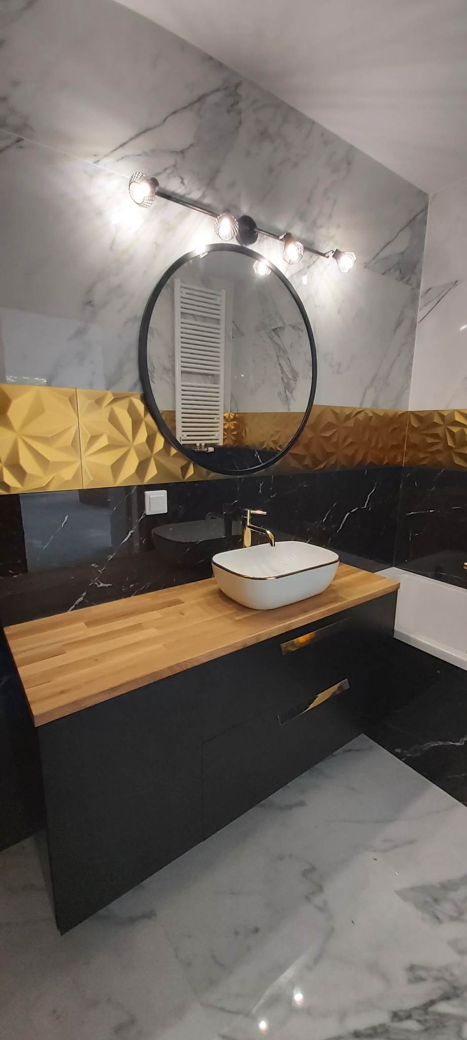 Nowoczesna łazienka z marmurowymi płytkami, złotymi akcentami i drewnianym blatem pod umywalką nablatową, oświetlona lampą z kilkoma żarówkami i okrągłym lustrem.