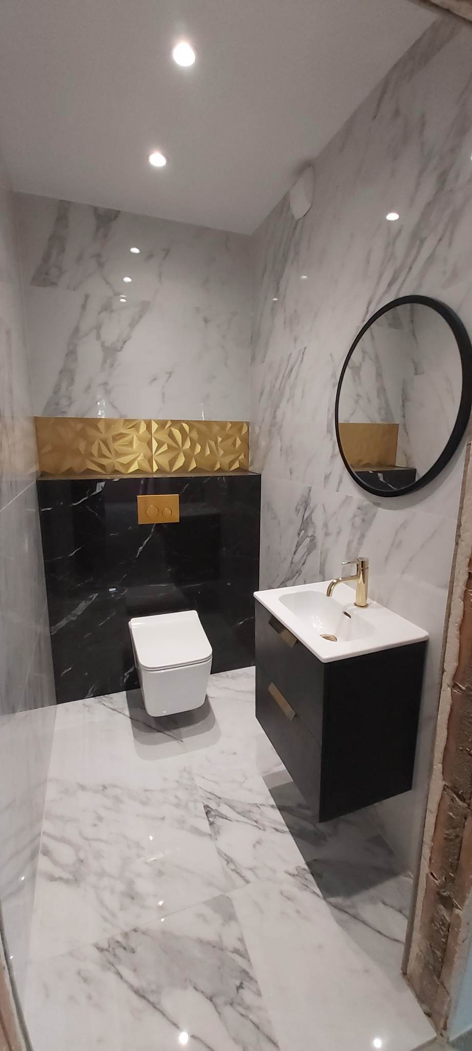 Nowoczesna łazienka z białymi marmurowymi płytkami, czarną zabudową toalety z geometryczną złotą dekoracją, czarną szafką umywalkową z okrągłym lustrem i złotą armaturą.