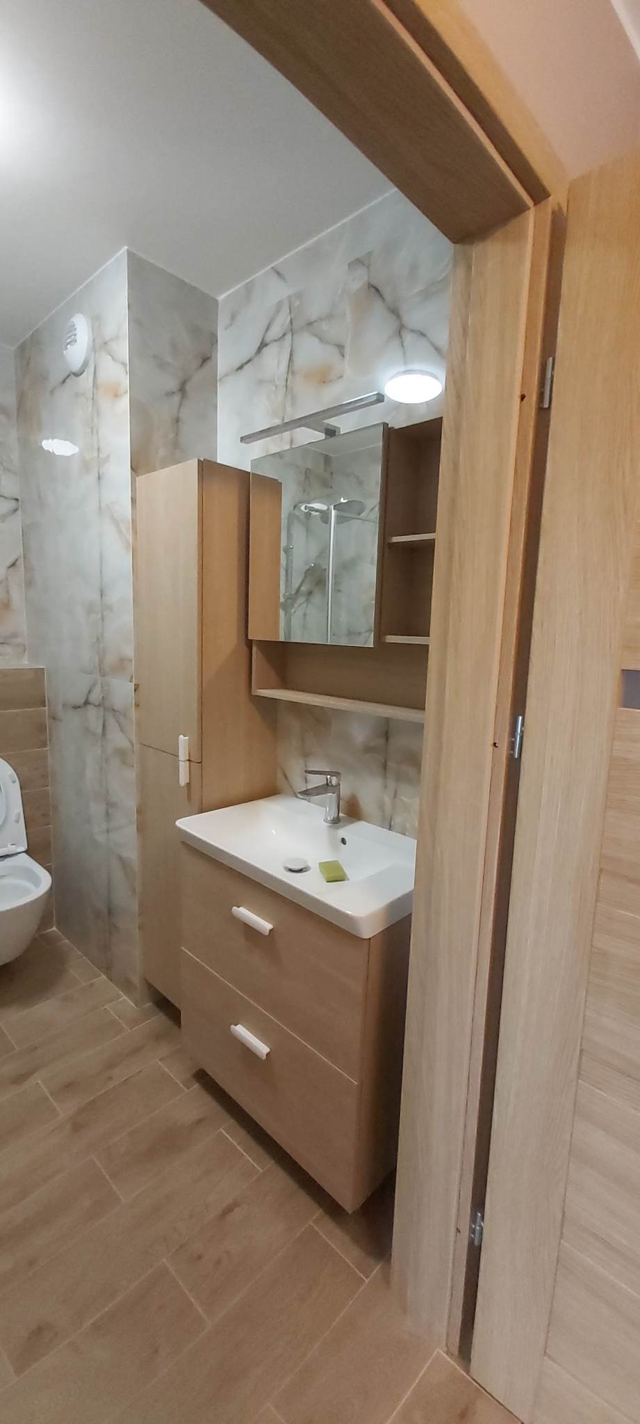 Wykończona łazienka z marmurowymi płytkami ściennymi, drewnopodobną podłogą, szafką z umywalką i lustrem z półkami, widok na kabinę prysznicową w lustrze.