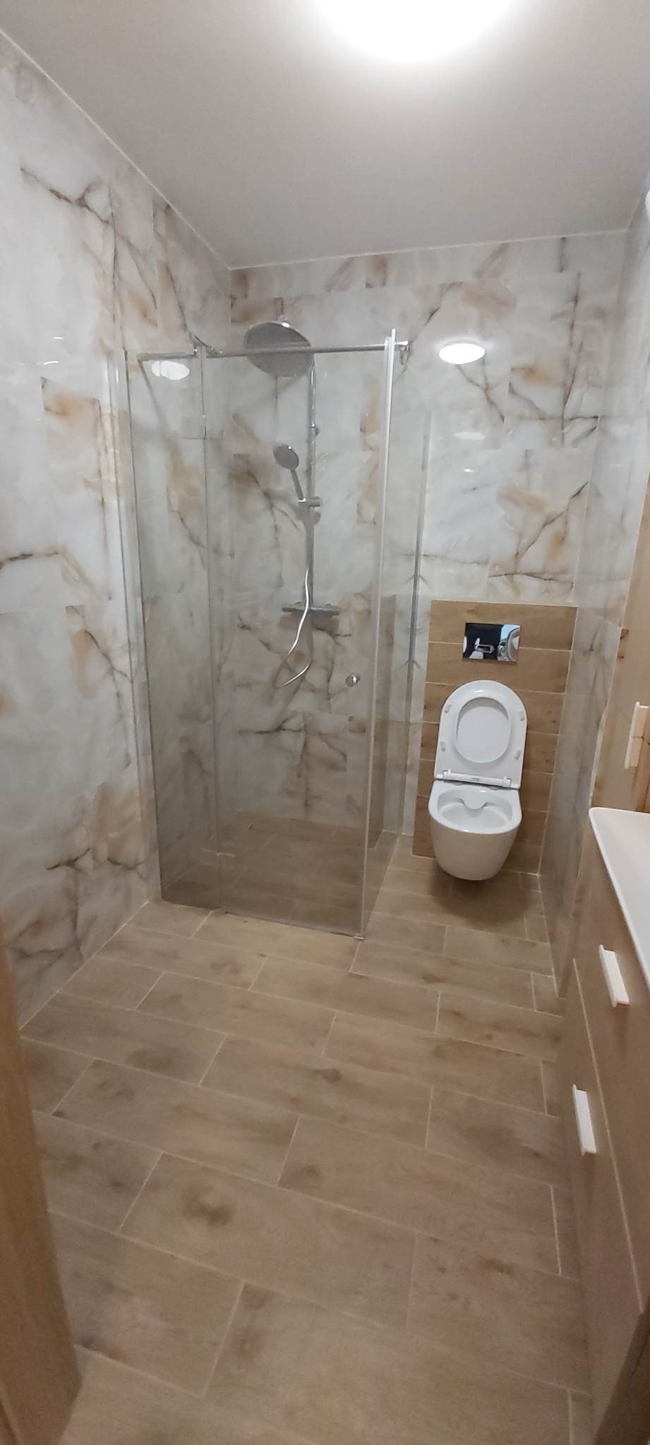 Nowoczesna łazienka po remoncie z kabiną prysznicową ze szkła, wiszącą toaletą i płytkami imitującymi marmur na ścianach oraz drewnopodobnymi na podłodze.