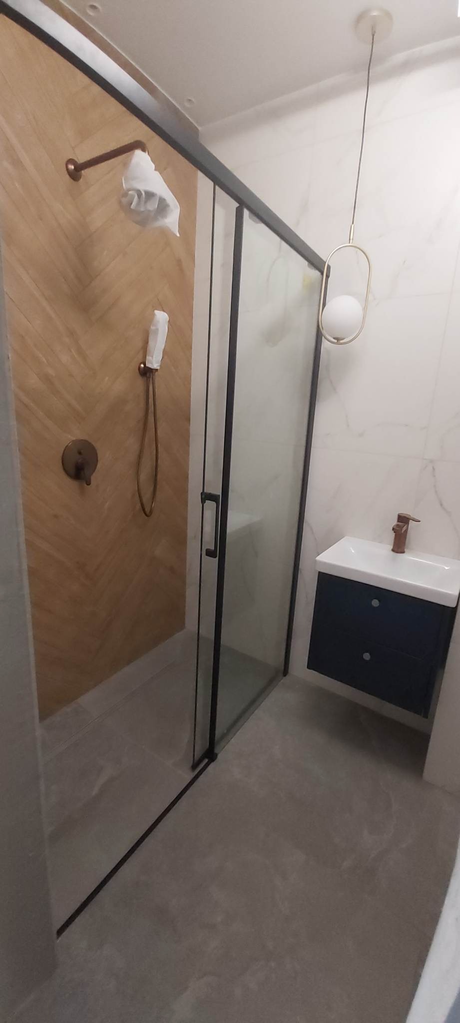 Nowoczesna łazienka z kabiną prysznicową z czarnymi ramami, drewnopodobnymi płytkami na ścianie prysznica, armaturą w kolorze miedzi, wiszącą szafką z umywalką i designerską lampą sufitową.