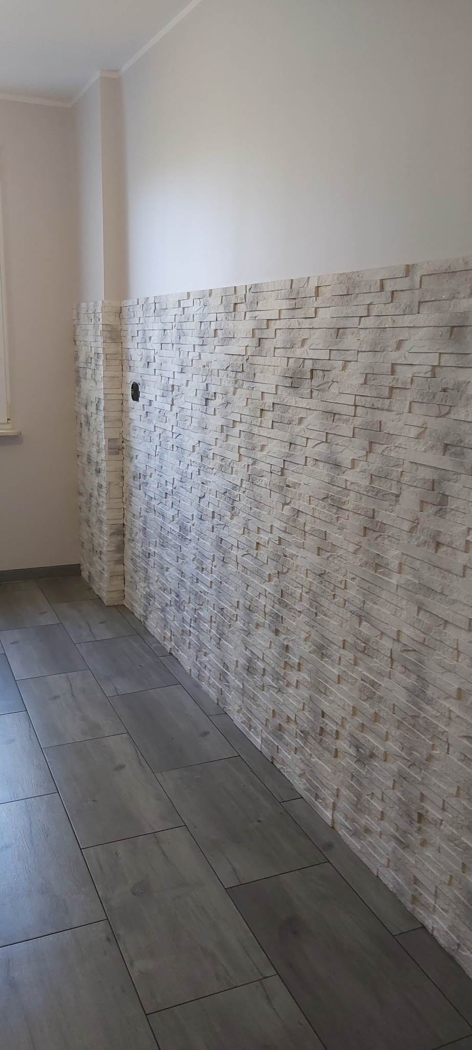Ściana wyłożona dekoracyjną, jasną imitacją kamienia, z widocznym otworem na gniazdko elektryczne, połączona z szarą podłogą z płytek imitujących drewno.