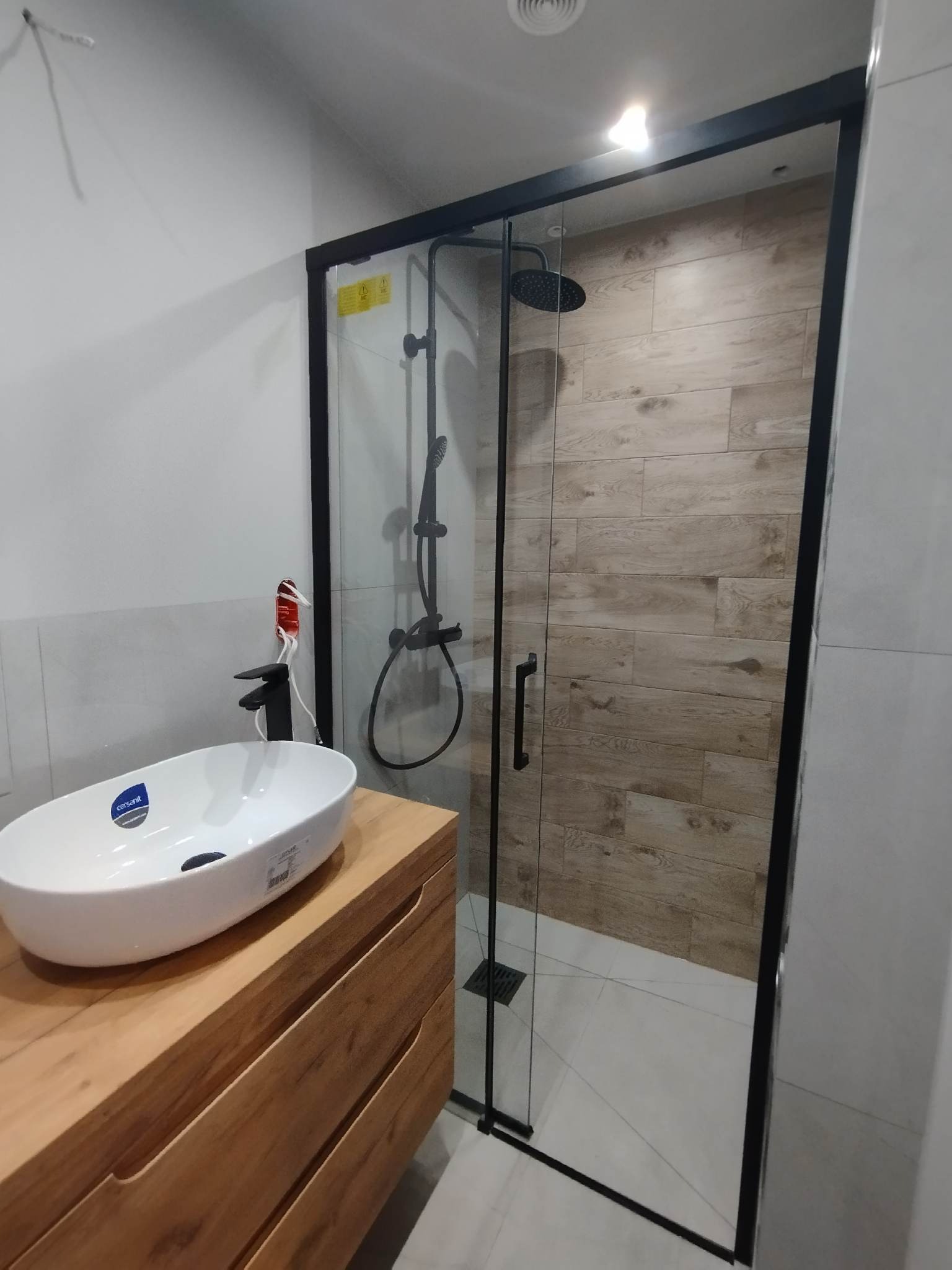 Nowoczesna łazienka w Rumi z kabiną prysznicową typu walk-in z czarną ramą i deszczownicą, umywalką nablatową na drewnianej szafce oraz płytkami imitującymi drewno na ścianie w kabinie.