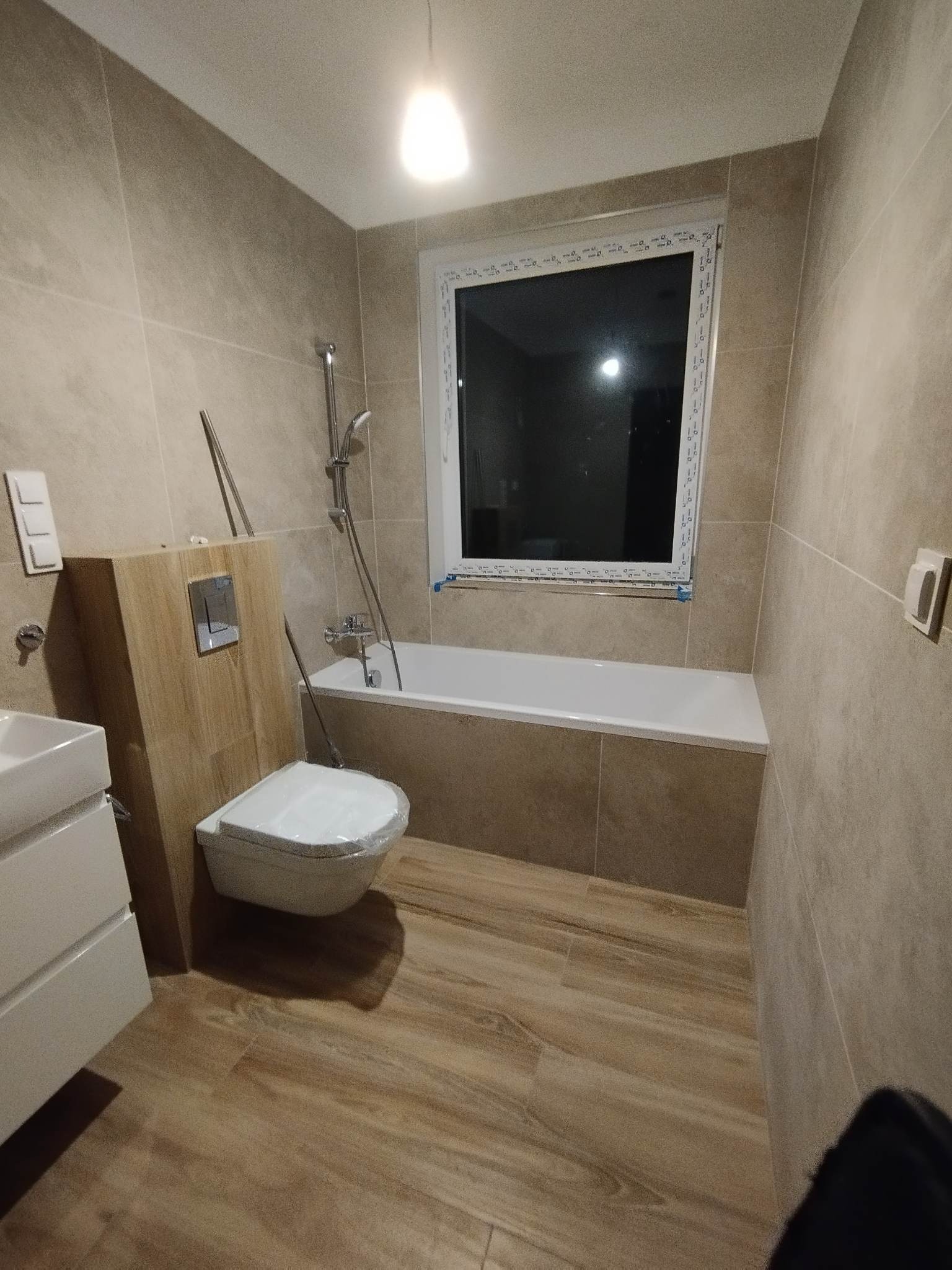 Nowoczesna łazienka w beżowych odcieniach z drewnopodobną podłogą i płytkami imitującymi kamień na ścianach, z białą wanną, umywalką z szafką i toaletą jeszcze w folii ochronnej.