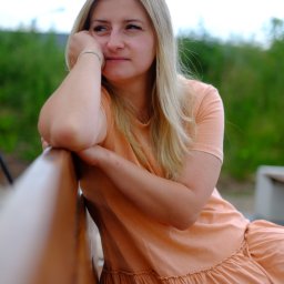 DamanFoto - Portret kobiety z blond włosami w brzoskwiniowej sukience, opierającej się o drewniane oparcie ławki na tle zieleni. Spokojny wyraz twarzy, naturalne światło.