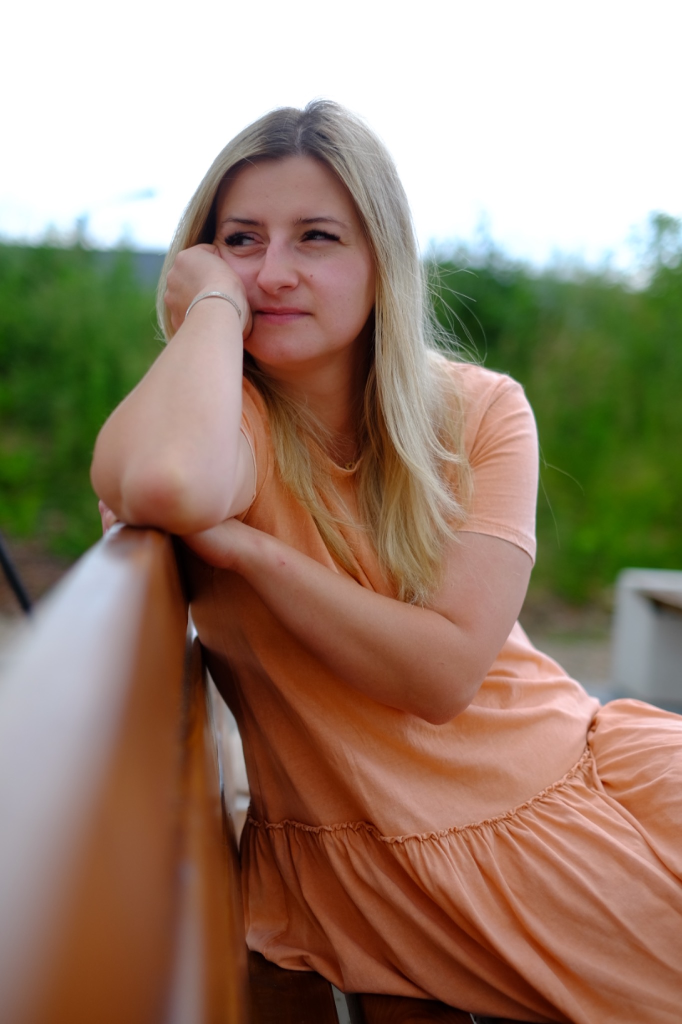 Portret kobiety z blond włosami w brzoskwiniowej sukience, opierającej się o drewniane oparcie ławki na tle zieleni. Spokojny wyraz twarzy, naturalne światło.