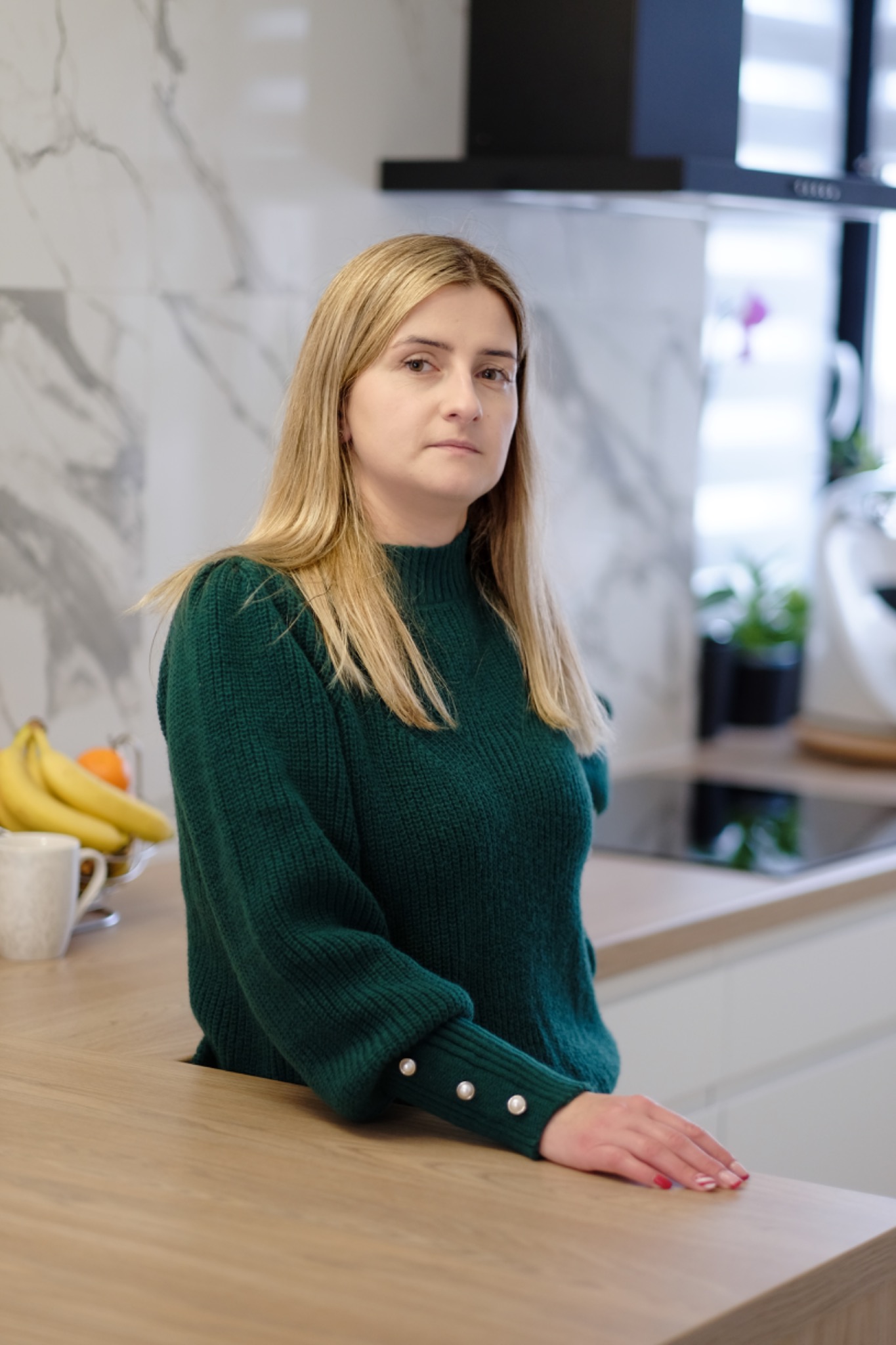 Portret kobiety w zielonym swetrze z perłowymi guzikami na tle nowoczesnej kuchni z marmurową ścianą, owocami i roślinami.