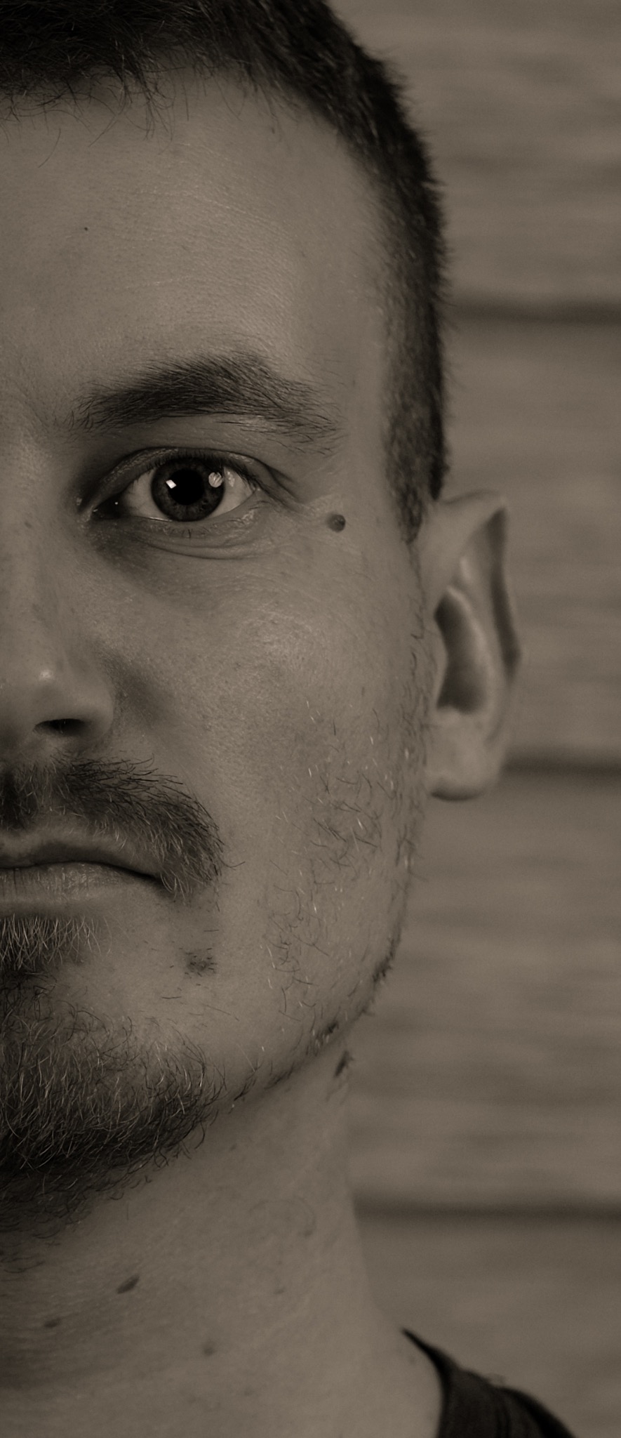 Portret w sepii, zbliżenie na połowę twarzy mężczyzny z zarostem, widoczne pieprzyki i faktura skóry na tle drewnianej ściany.