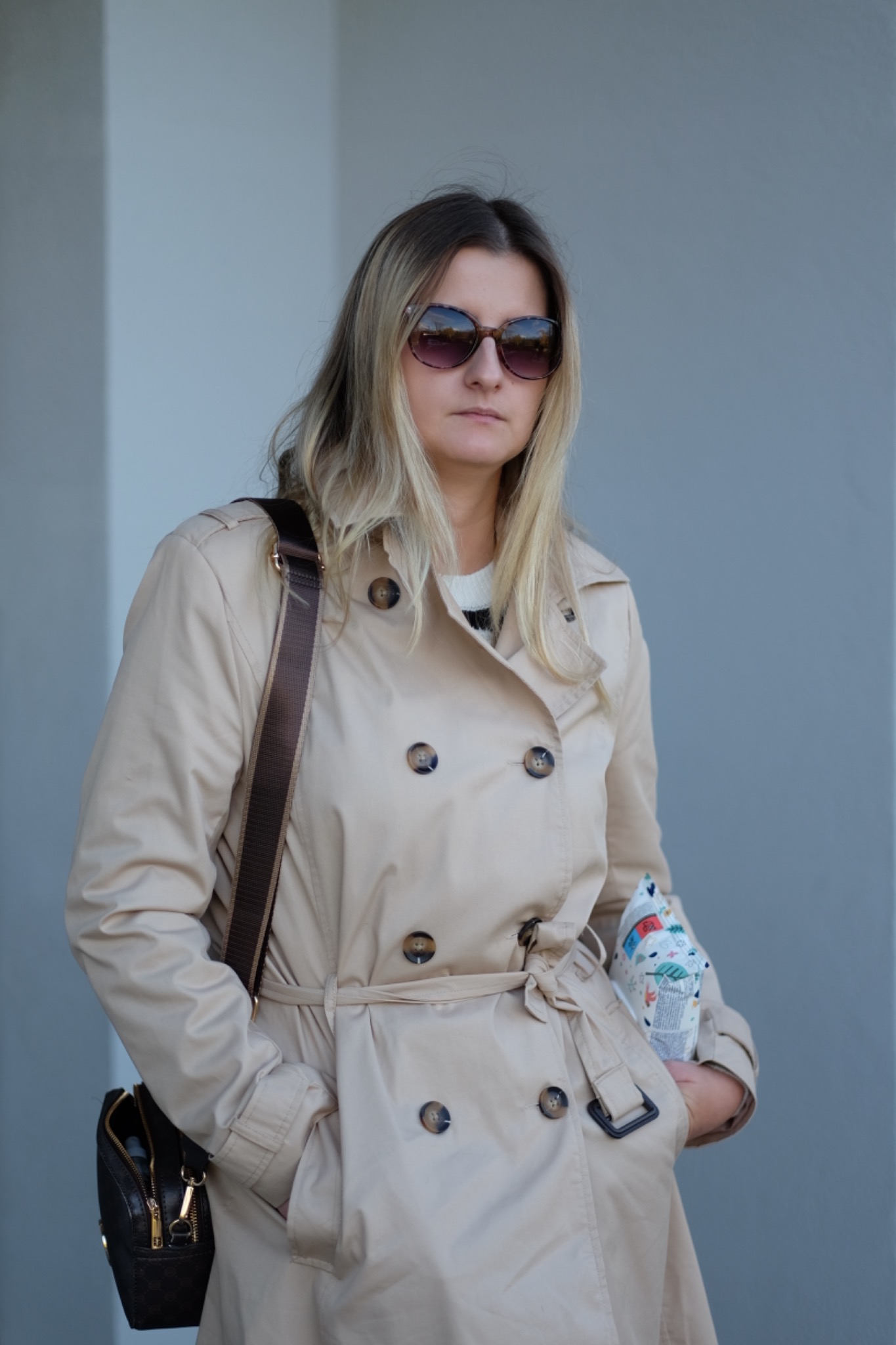 Portret kobiety w beżowym płaszczu, okularach przeciwsłonecznych i z torbą na ramieniu, trzymającej w ręku kolorową torbę na zakupy na tle jasnoszarej ściany.