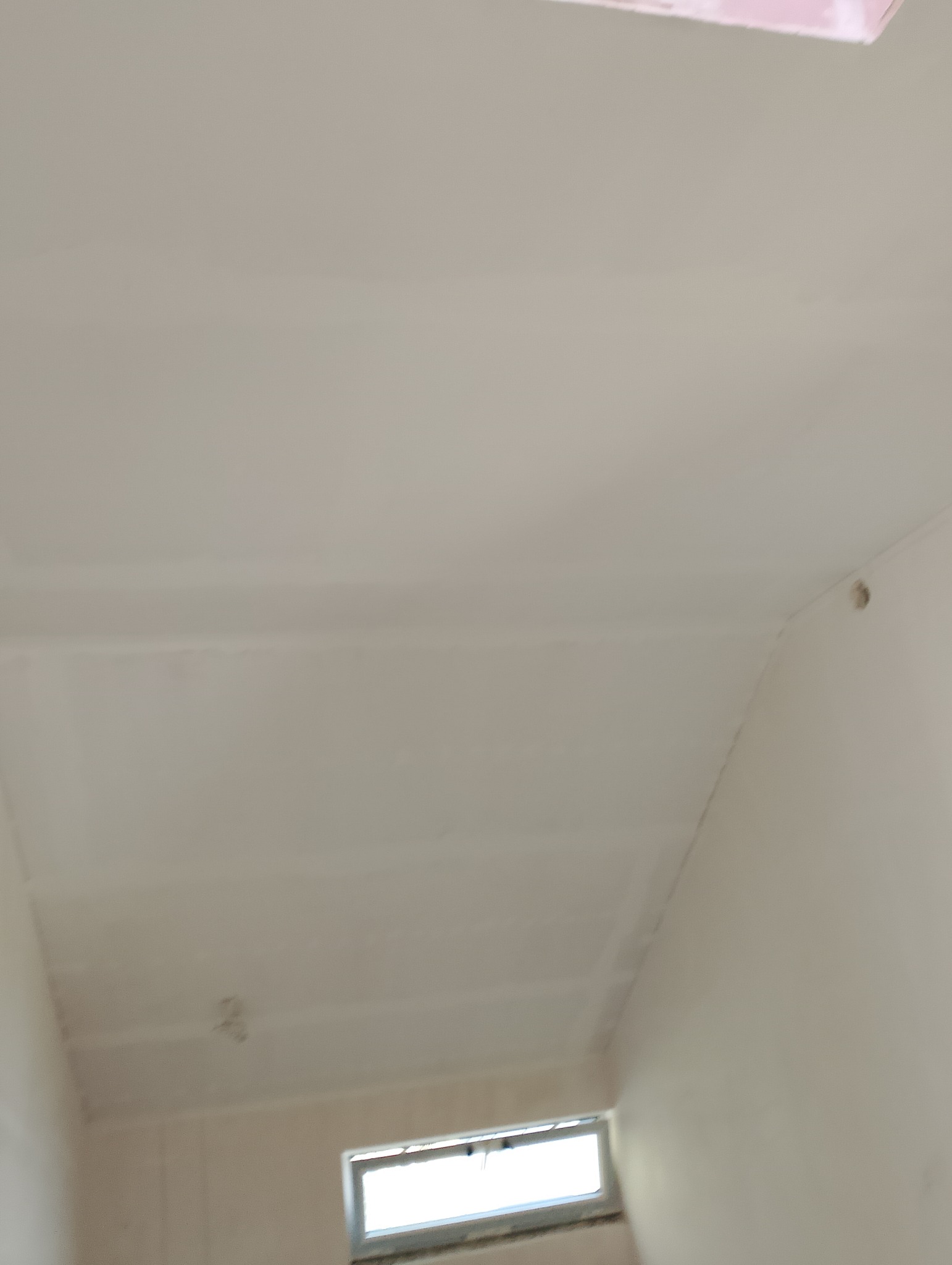 Biały sufit po szpachlowaniu, widoczne niedoskonałości i łączenia płyt kartonowo-gipsowych, przygotowany do dalszych prac wykończeniowych, małe okno w tle.