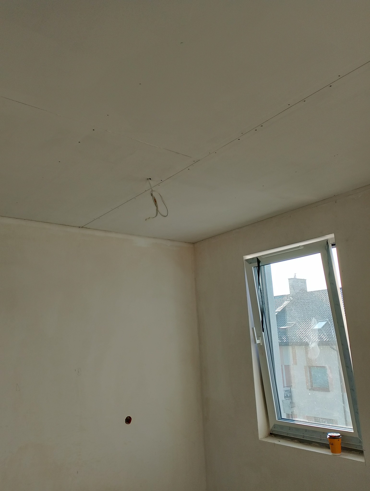 Pokój w trakcie remontu: ściany przygotowane do malowania, widoczne otwory po kołkach na suficie z zamontowaną płytą, okno dachowe z kubkiem po kawie na parapecie.